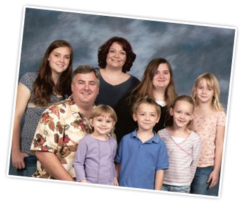 Family portrait of seven people including adults and children, smiling against a gray background