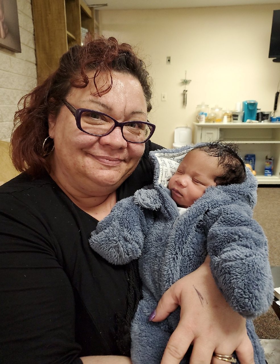 A woman with glasses and earrings smiles as she holds a newborn baby wrapped in a gray fleece blanket. The background shows a kitchen shelf with various items.