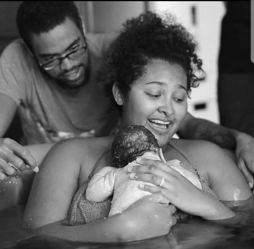 A woman breastfeeds a newborn baby while her partner leans over them and kisses her forehead.