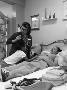 A woman with glasses and curly hair holding a medical instrument and talking to a young girl sitting on a hospital bed next to a girl lying in bed with her eyes closed, in a hospital room with light green walls and decorative shelves.