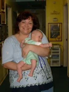 A woman with curly brown hair smiling and holding a baby in a hospital or medical setting.