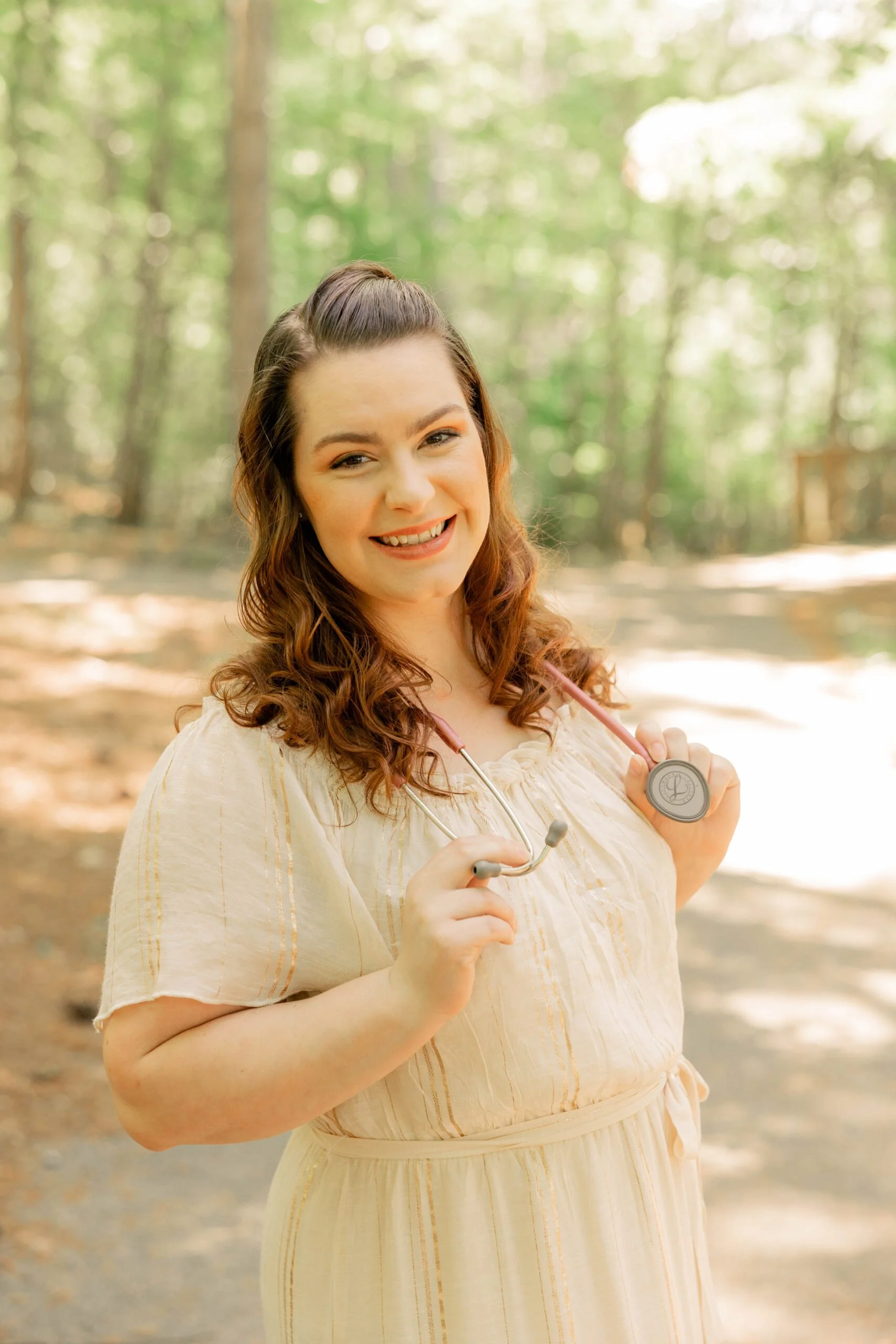 A smiling woman with long curly hair holding a stethoscope in a forest setting.