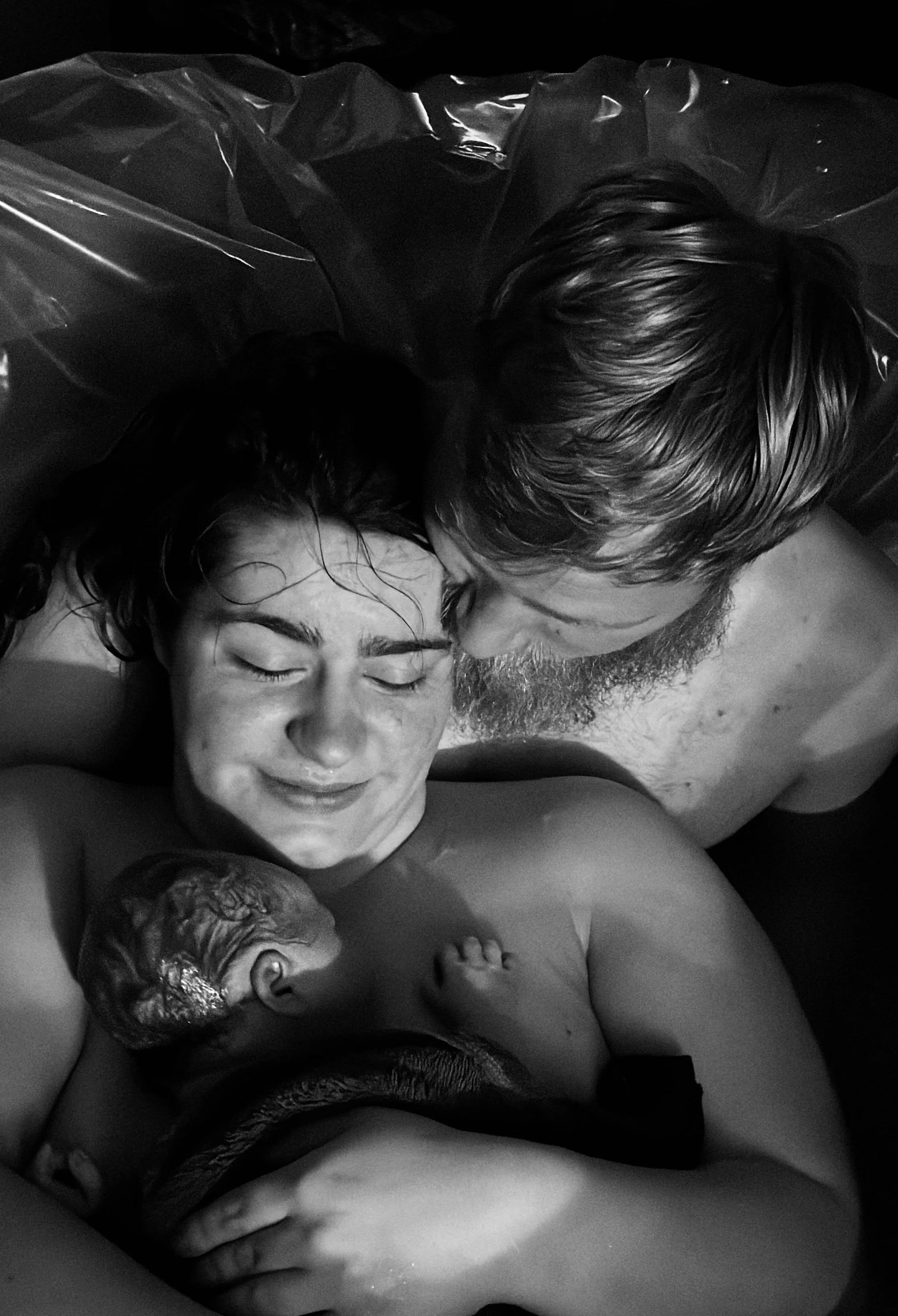 A woman breastfeeds a newborn baby while her partner leans over them and kisses her forehead.