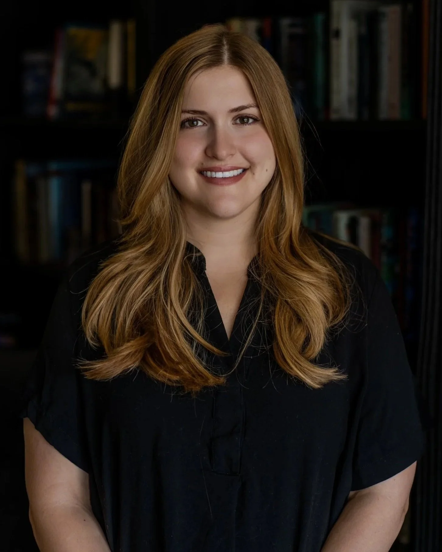 Portrait of a young woman with long, wavy brown hair, wearing a black top, standing in front of a dark bookshelf filled with books.