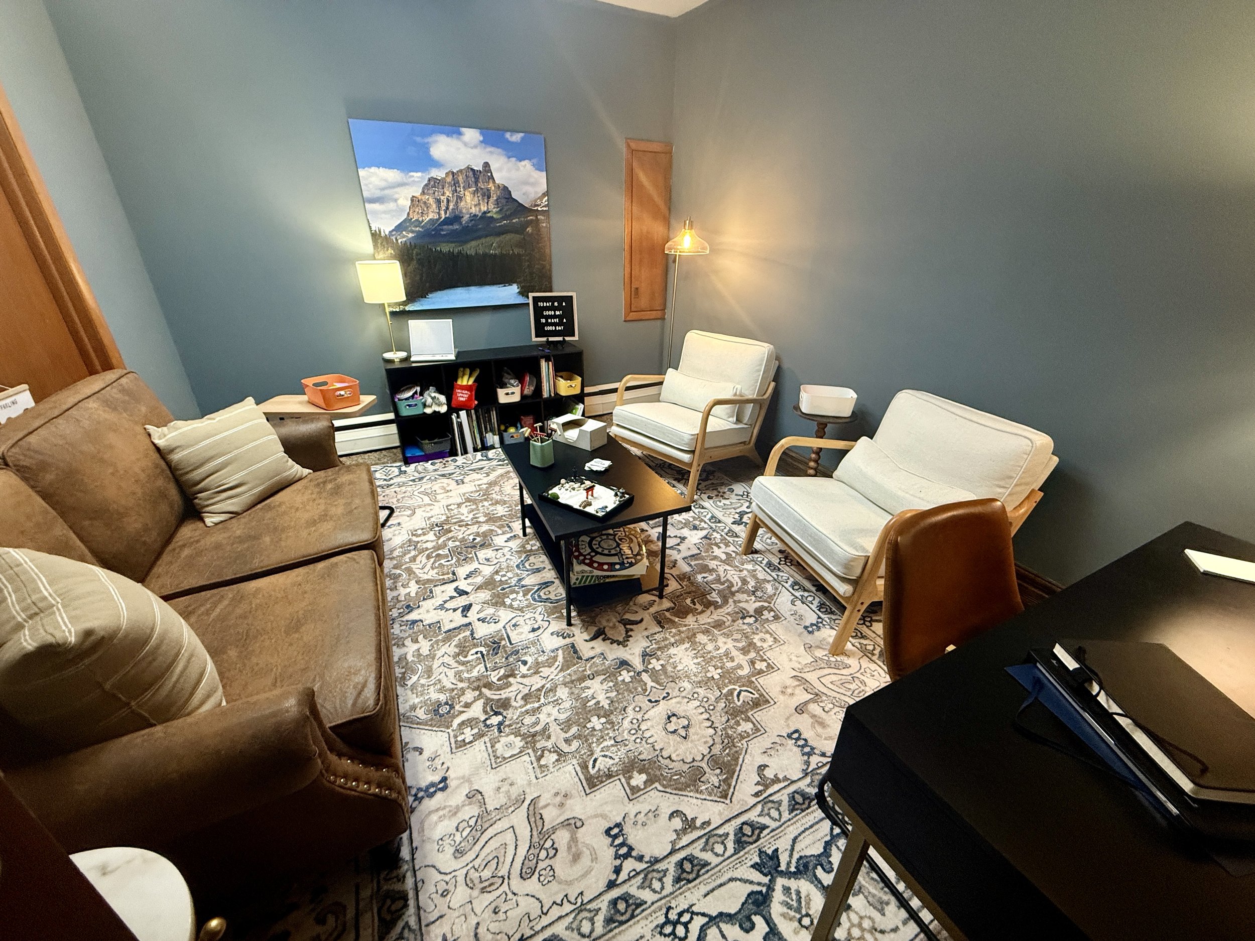 Living room with a brown sofa, two white armchairs, a black coffee table, a patterned area rug, a small black bookshelf, a table lamp, a window, and various office items on a black desk.