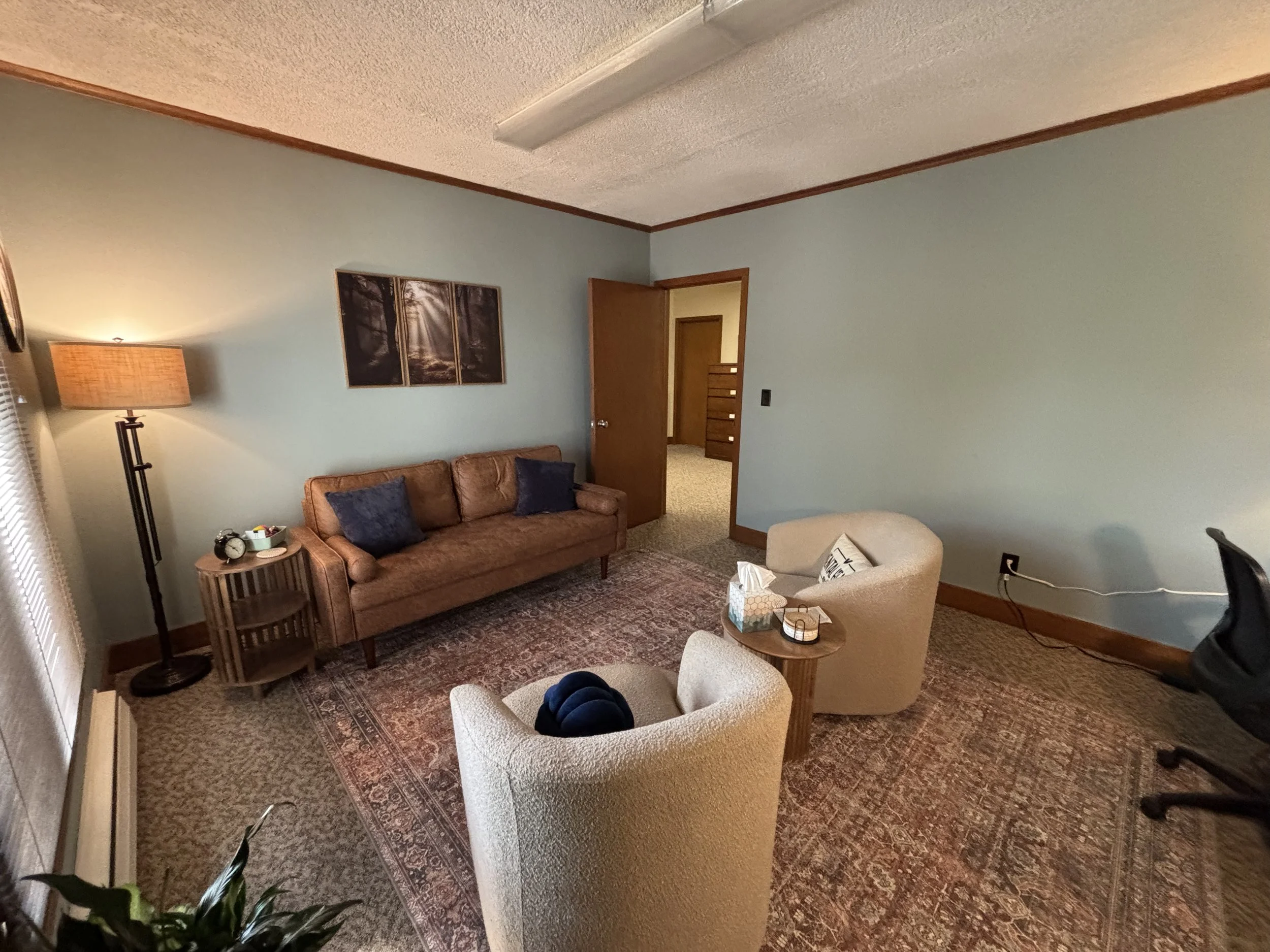 Living room with light green walls, brown couch with two navy pillows, two cream armchairs, a wooden side table, a floor lamp, a patterned rug, a painting of a forest on the wall, and an open doorway leading to another room.