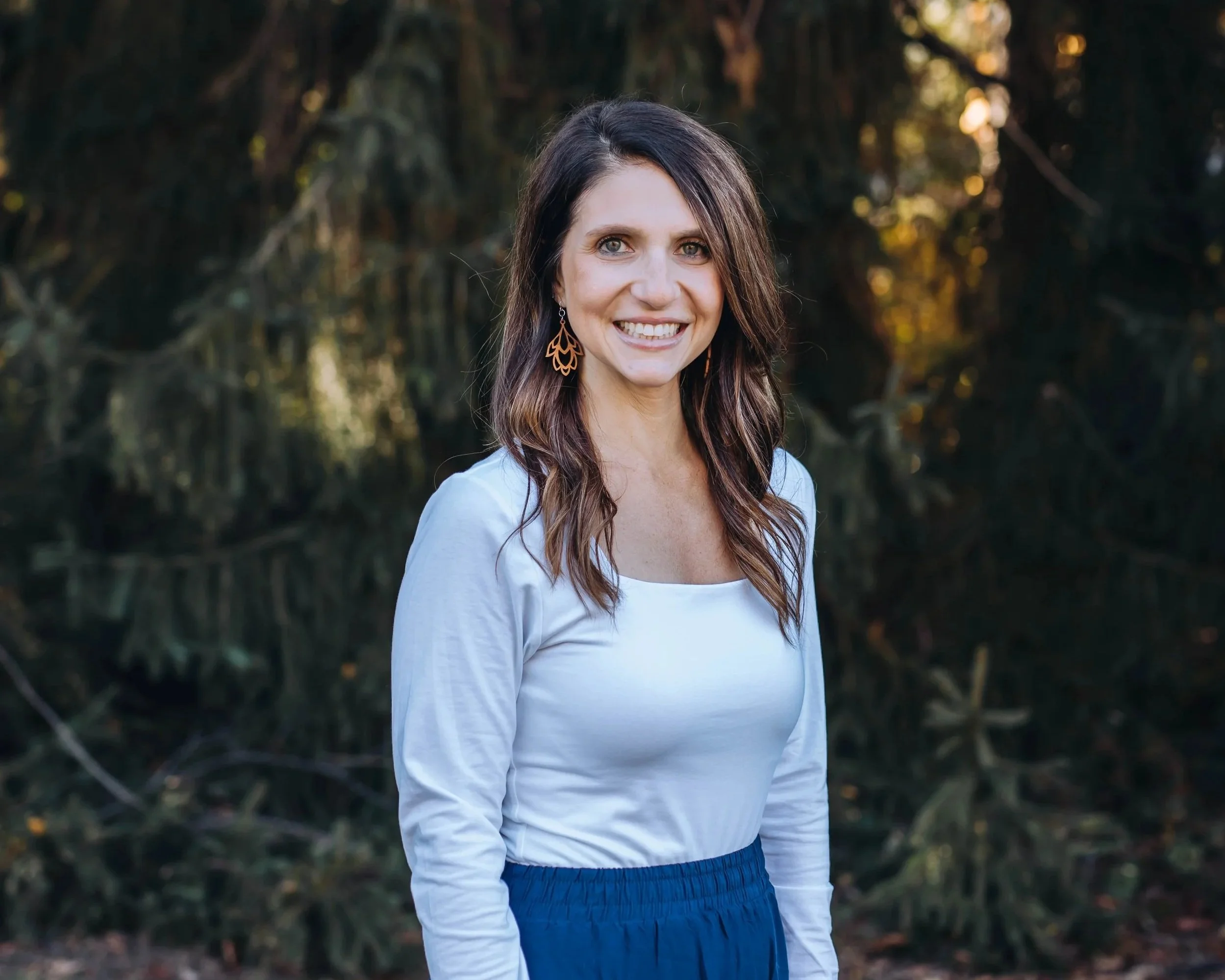 A woman with long, wavy brown hair smiling outdoors in front of a dark green, leafy background, wearing a white long-sleeve top and blue skirt, with orange earrings.