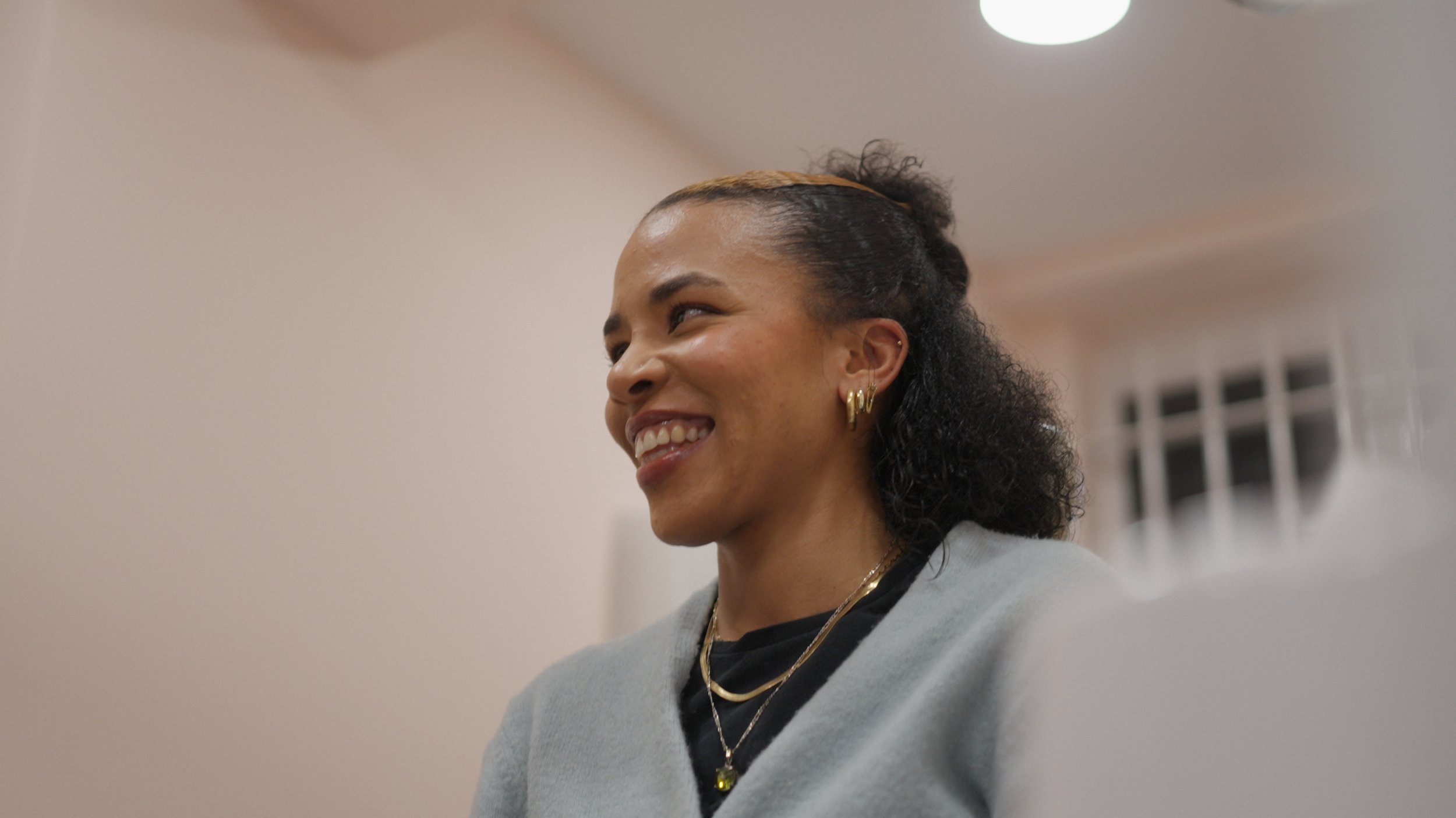 A smiling woman with dark curly hair tied back, wearing gold hoop earrings and layered necklaces, sitting indoors with a neutral-colored wall and ceiling light in the background.