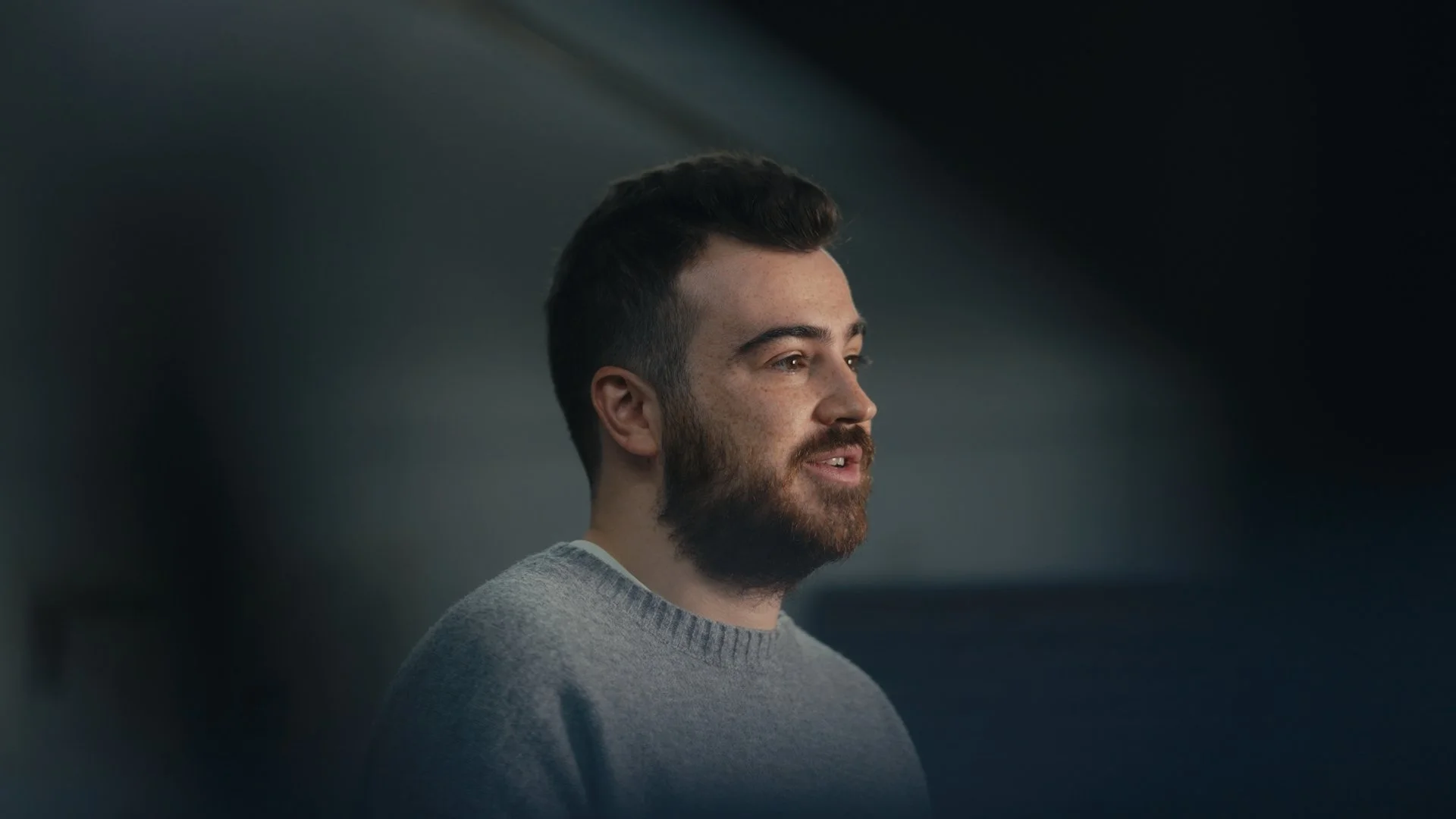 A young man with a beard and short dark hair, wearing a gray sweater, looking to the right, in a dimly lit indoor setting.
