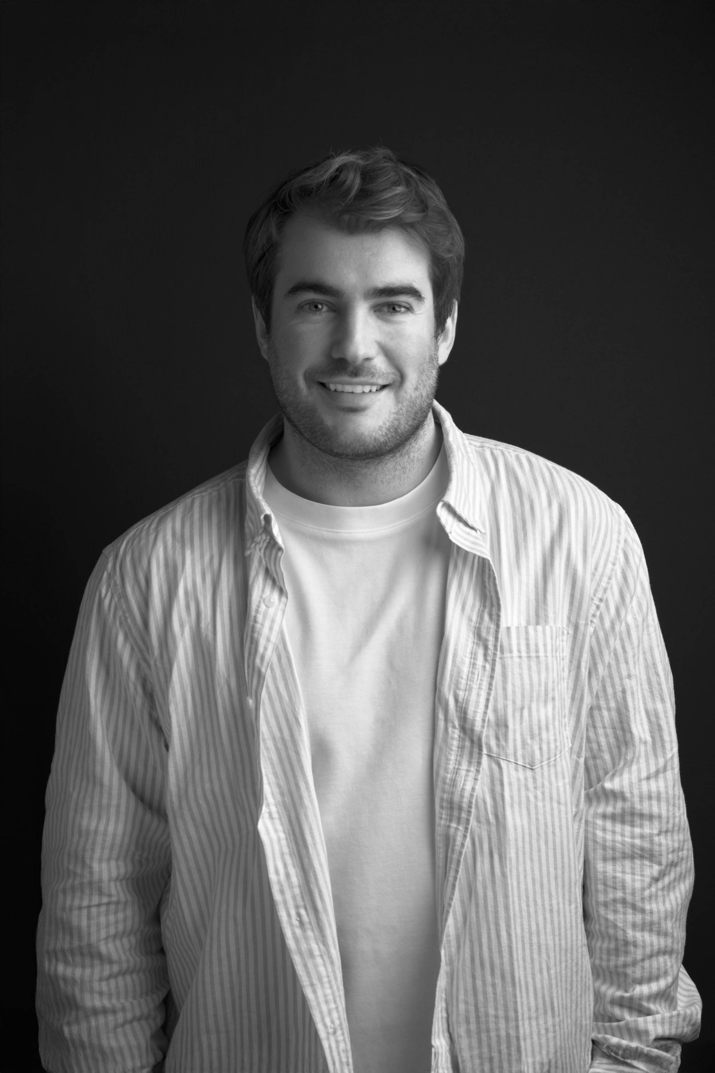 Black and white portrait of a young man with short hair, smiling, wearing a striped button-up shirt over a plain t-shirt, standing against a dark background.
