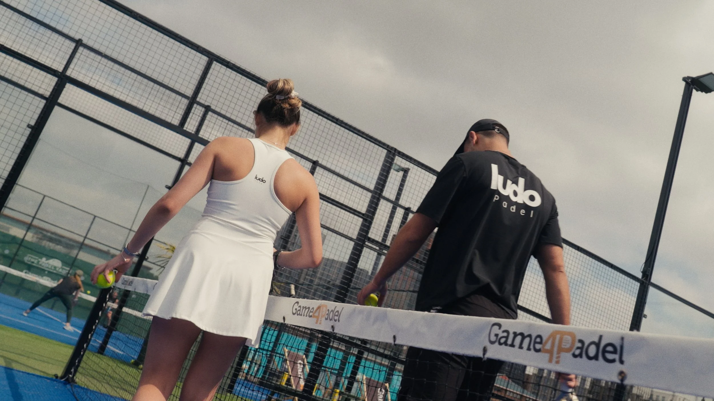 Two padel players, one woman and one man, walking away from the camera on a padel court, holding padel balls. The woman is wearing a white tennis dress, and the man is wearing a black shirt with 'Ludo Padel' written on the back. The court is surround