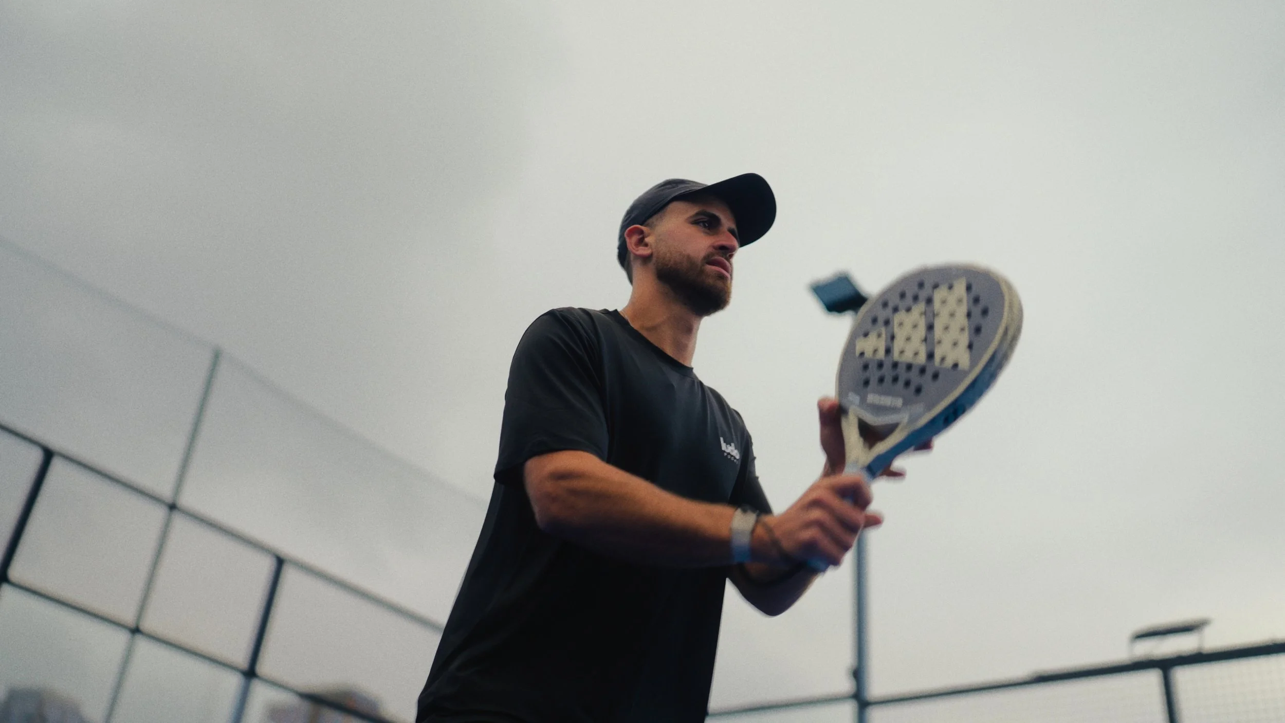 A man wearing a black cap and black t-shirt holding a tennis racket on an overcast day at the tennis court.