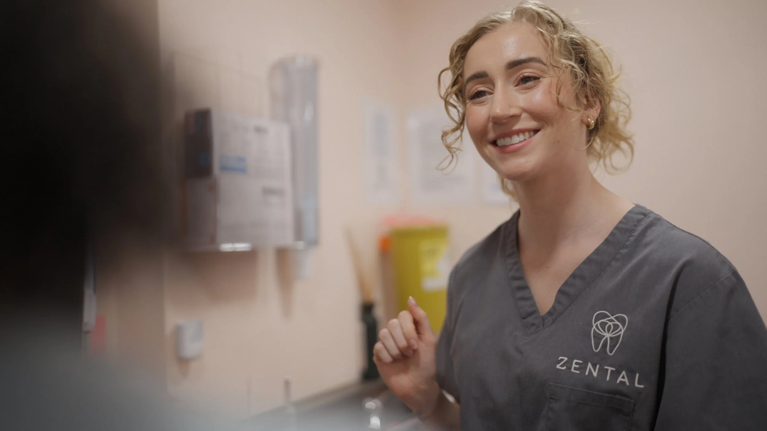 A smiling female dental professional with curly blonde hair wearing a gray scrubs with a dental logo and the word 'ZENTAL' on it, standing in a dental clinic treatment room.