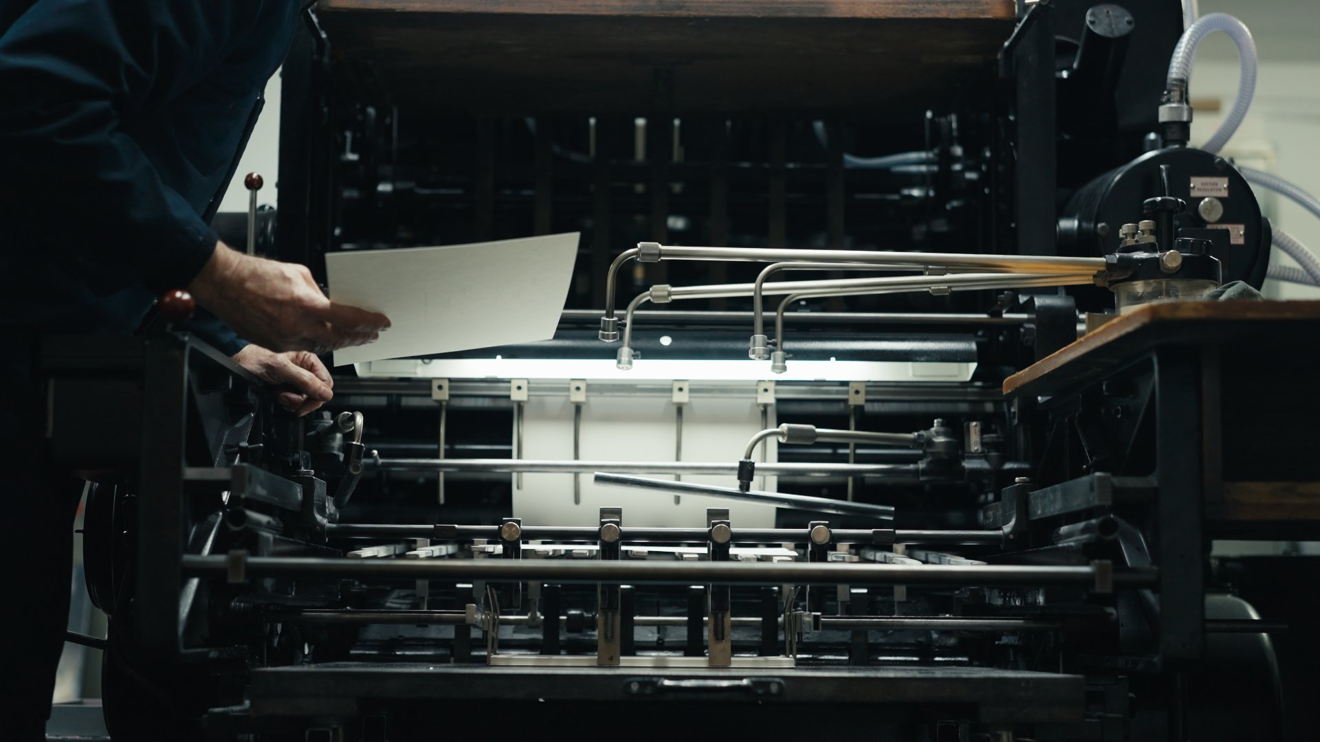 Close-up of an antique printing press, with a person placing or removing a sheet of paper, showing mechanical parts like metal arms and rollers.