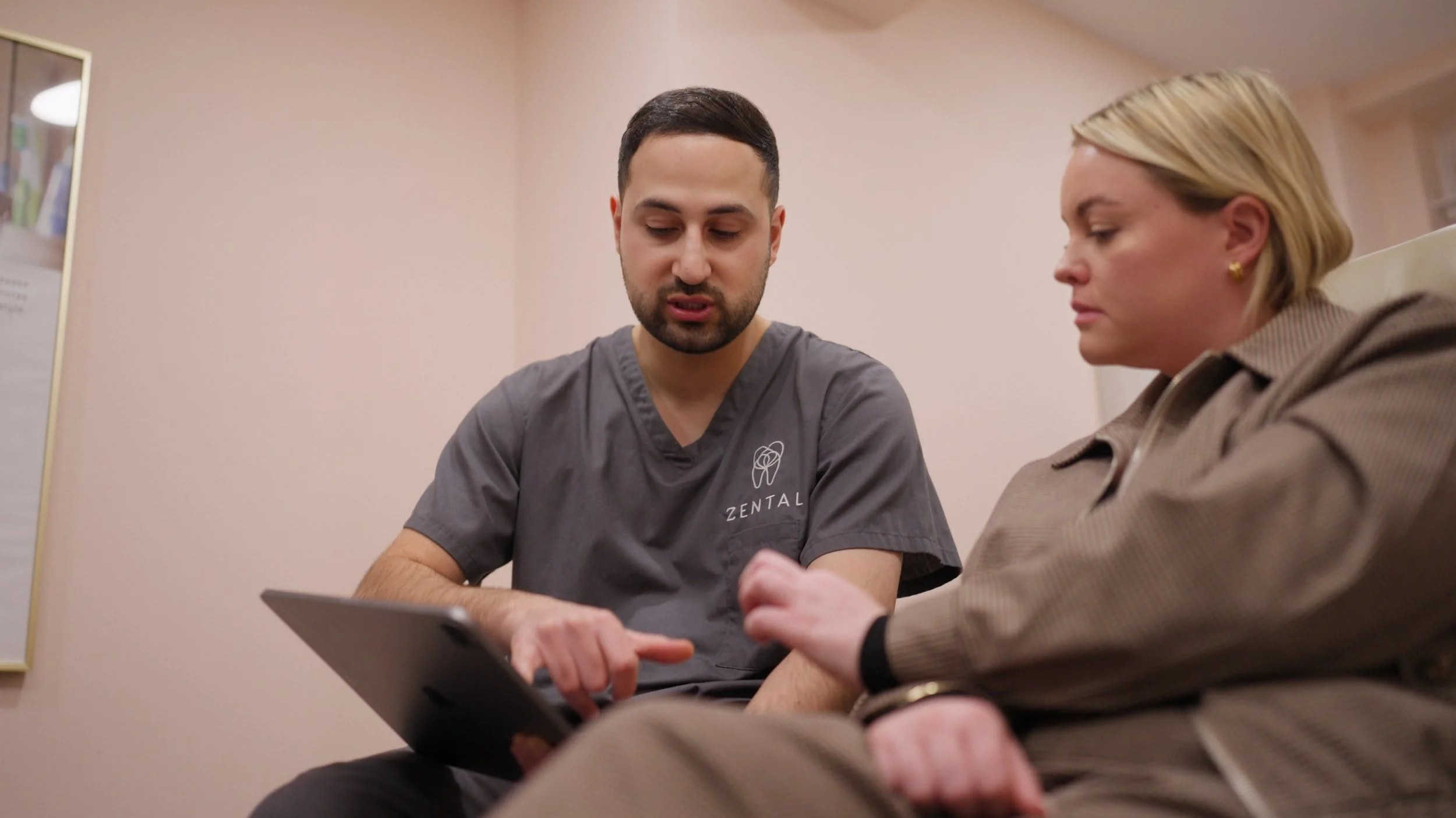 A male healthcare professional in gray scrubs with the logo 'Zental' on it, showing a woman in a beige jacket who is seated and listening.