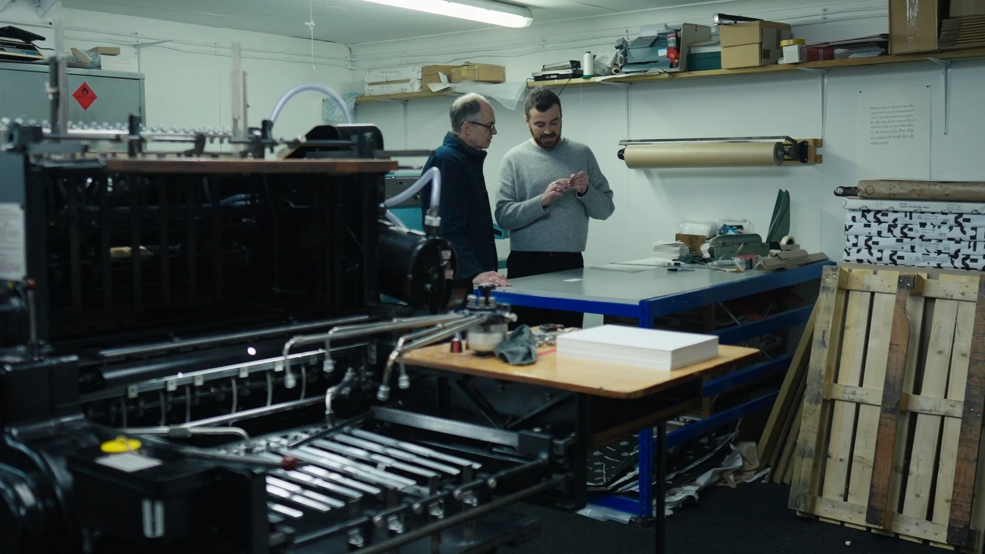 Two men working in a workshop surrounded by printing and packaging materials and equipment.