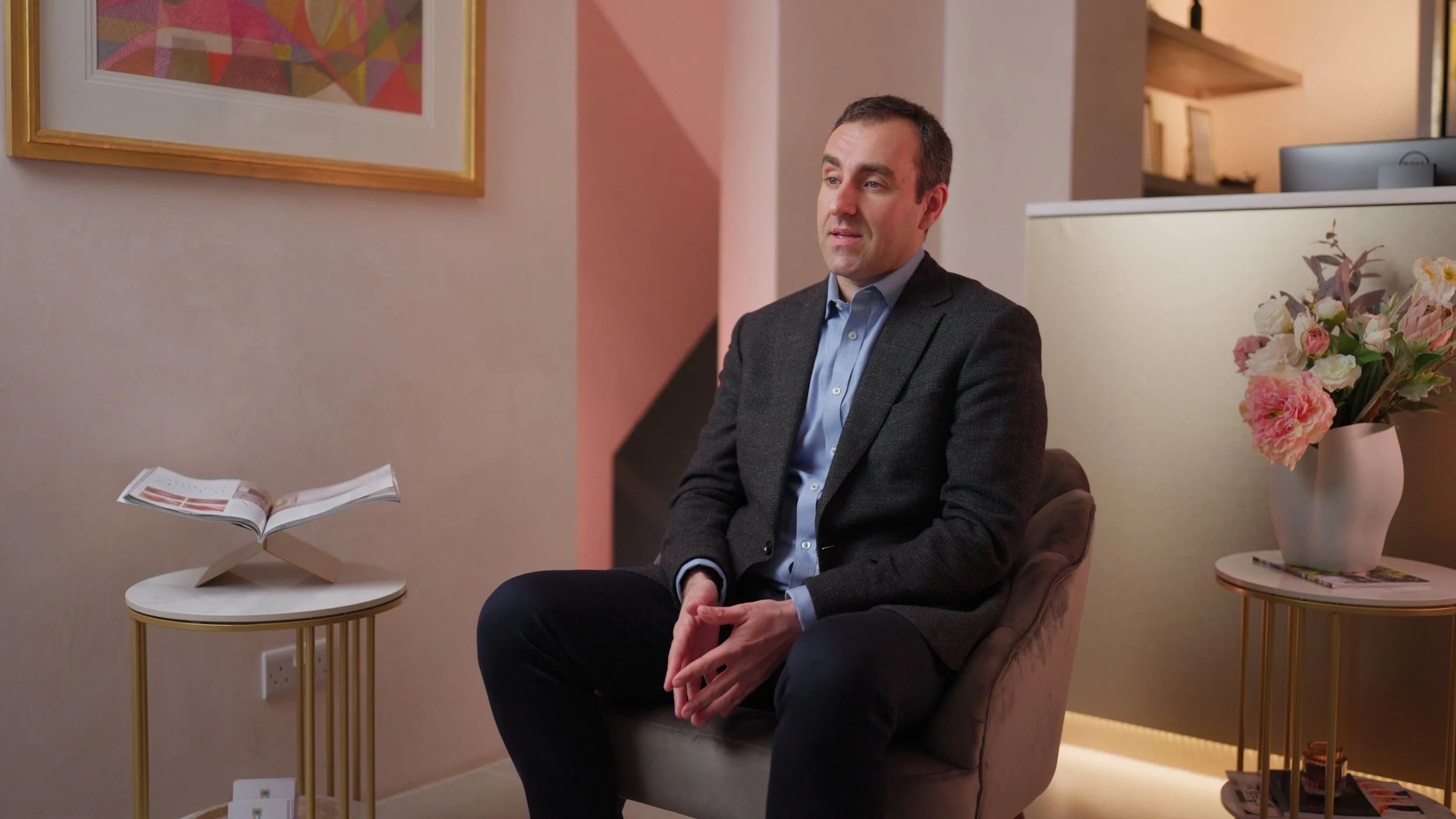 A man in a gray blazer sitting in a beige armchair in a room with pink and beige walls, low lighting, framed artwork, beside a small round table with an open magazine, and a larger table with a vase of pink and white flowers.