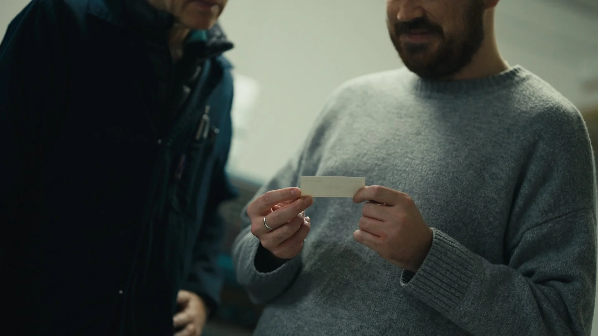 A man in a gray sweater holding a small card, standing next to a woman in dark clothing, in an indoor setting.