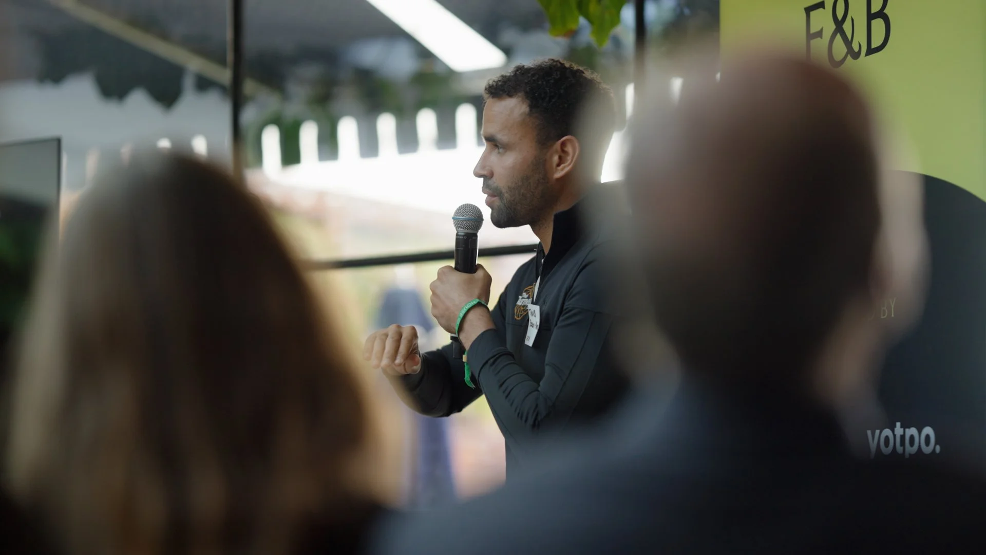Man holding a microphone, speaking at an event with people in the audience, in a tented outdoor setting.