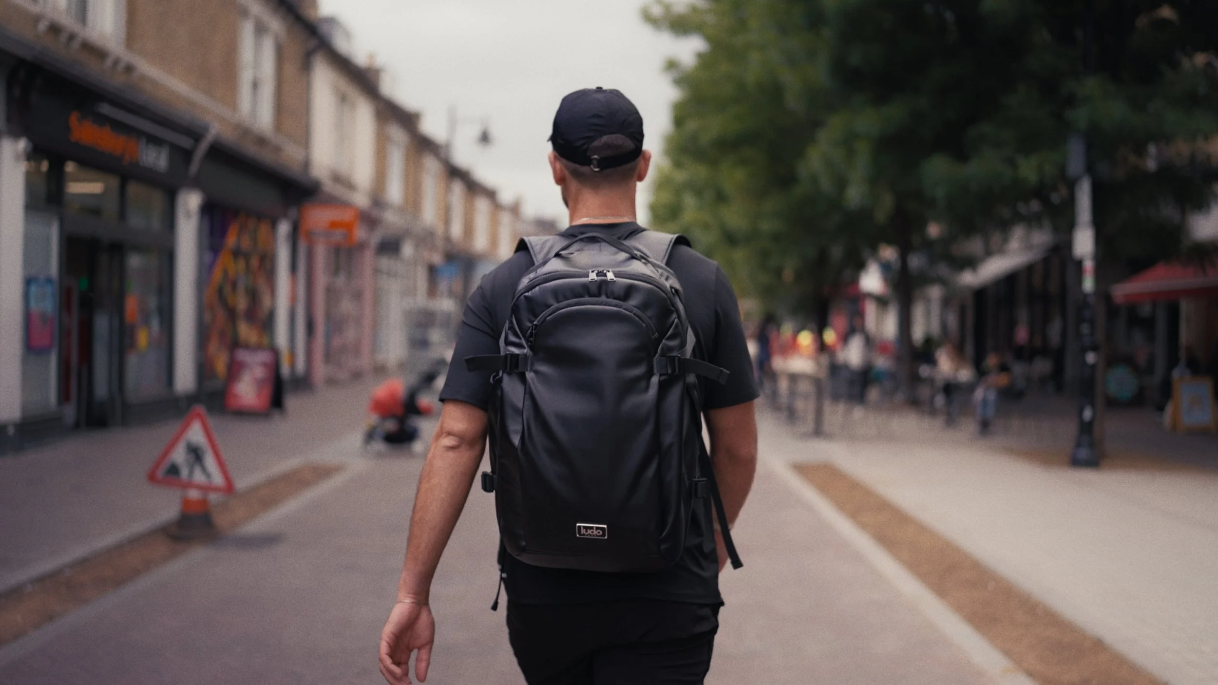 A person wearing a black cap, black t-shirt, and a black backpack walking down a city street with shops and trees, with people sitting at outdoor tables in the background.