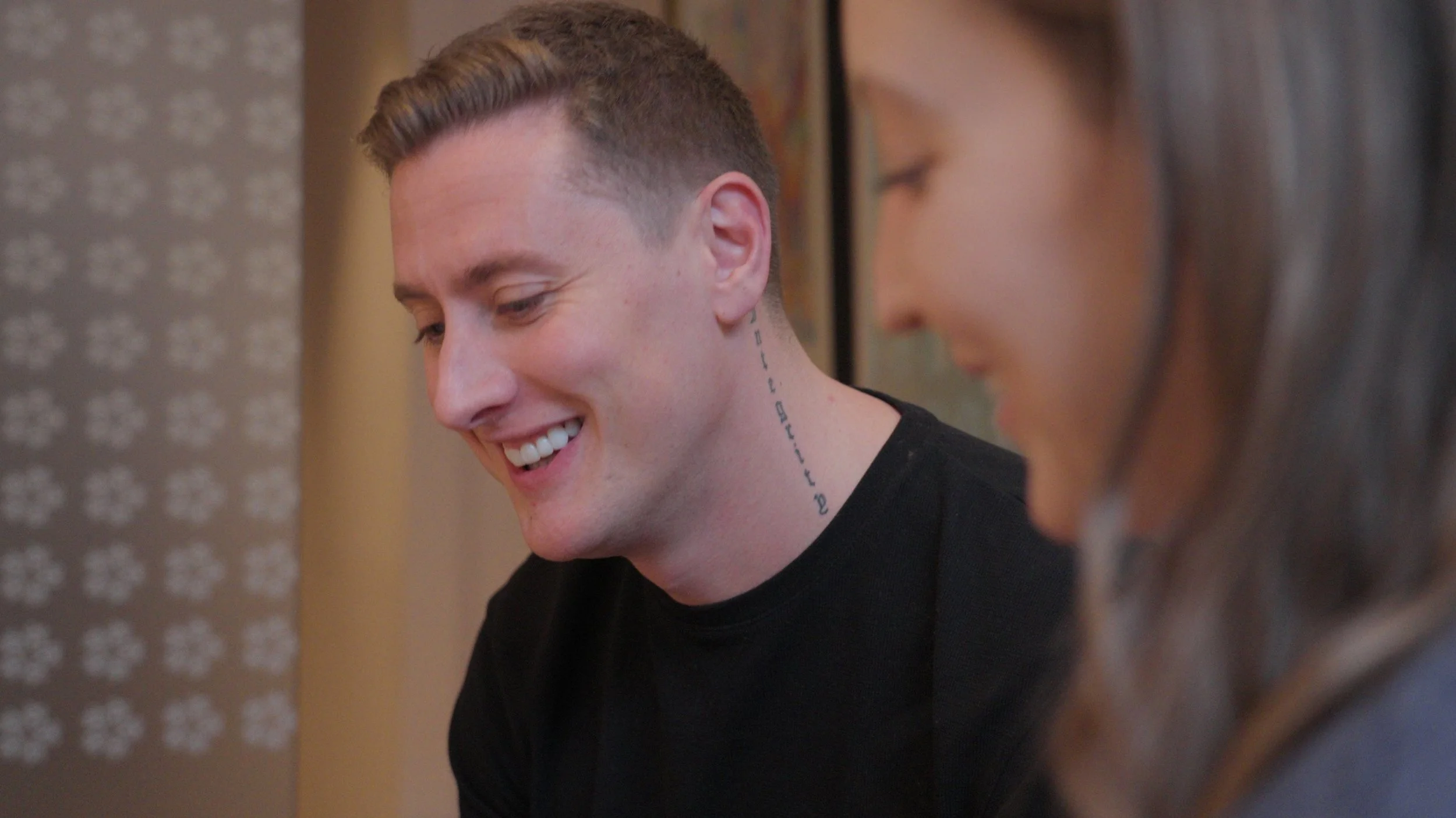 A smiling young man with short hair and a neck tattoo, and a woman with brown hair, sitting together indoors, looking downward.