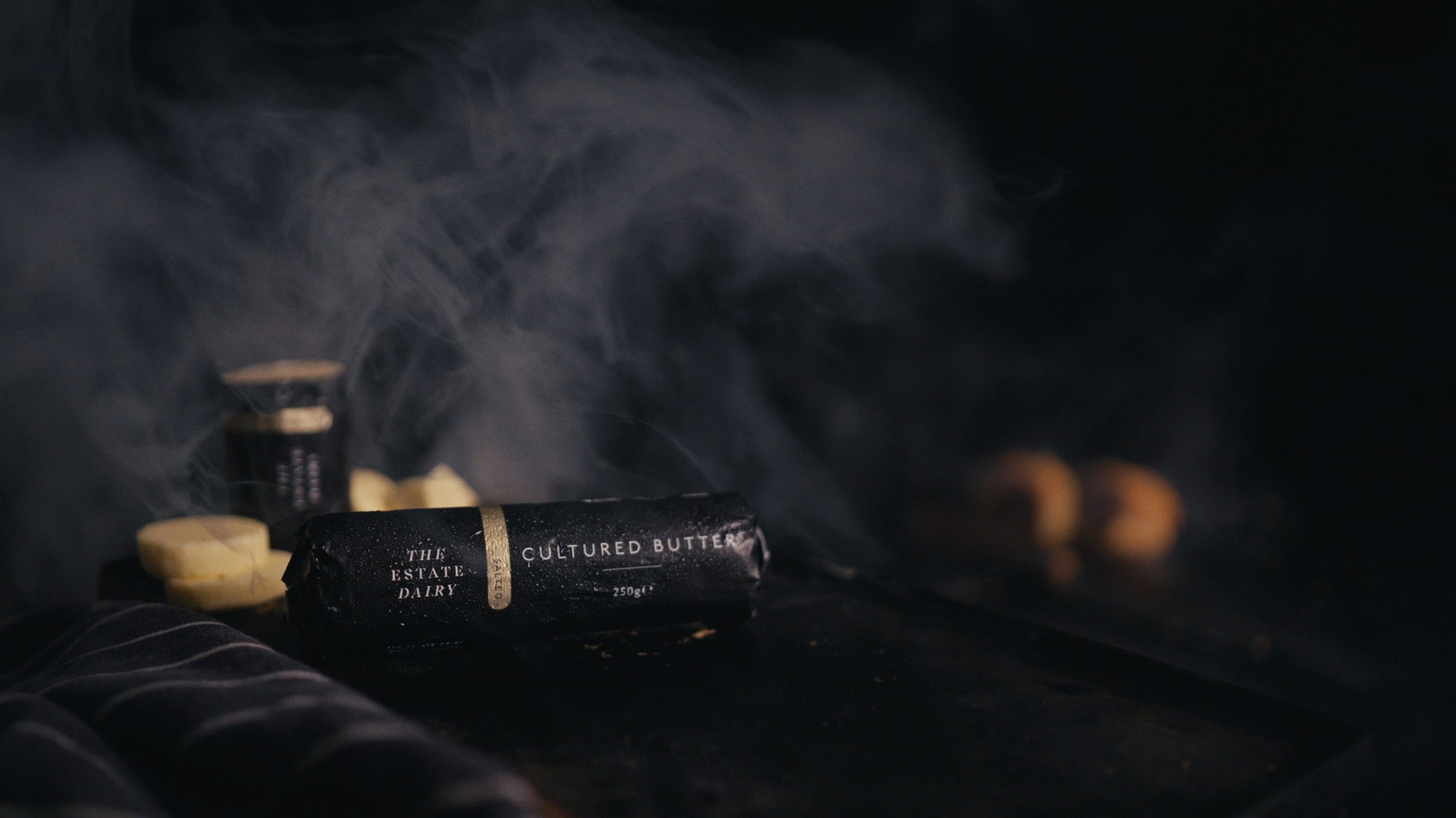 A black package labeled "Cultured Butter" with white and gold text, surrounded by several small, round cheese wheels and a small container, all on a dark surface with smoke or steam rising around them.