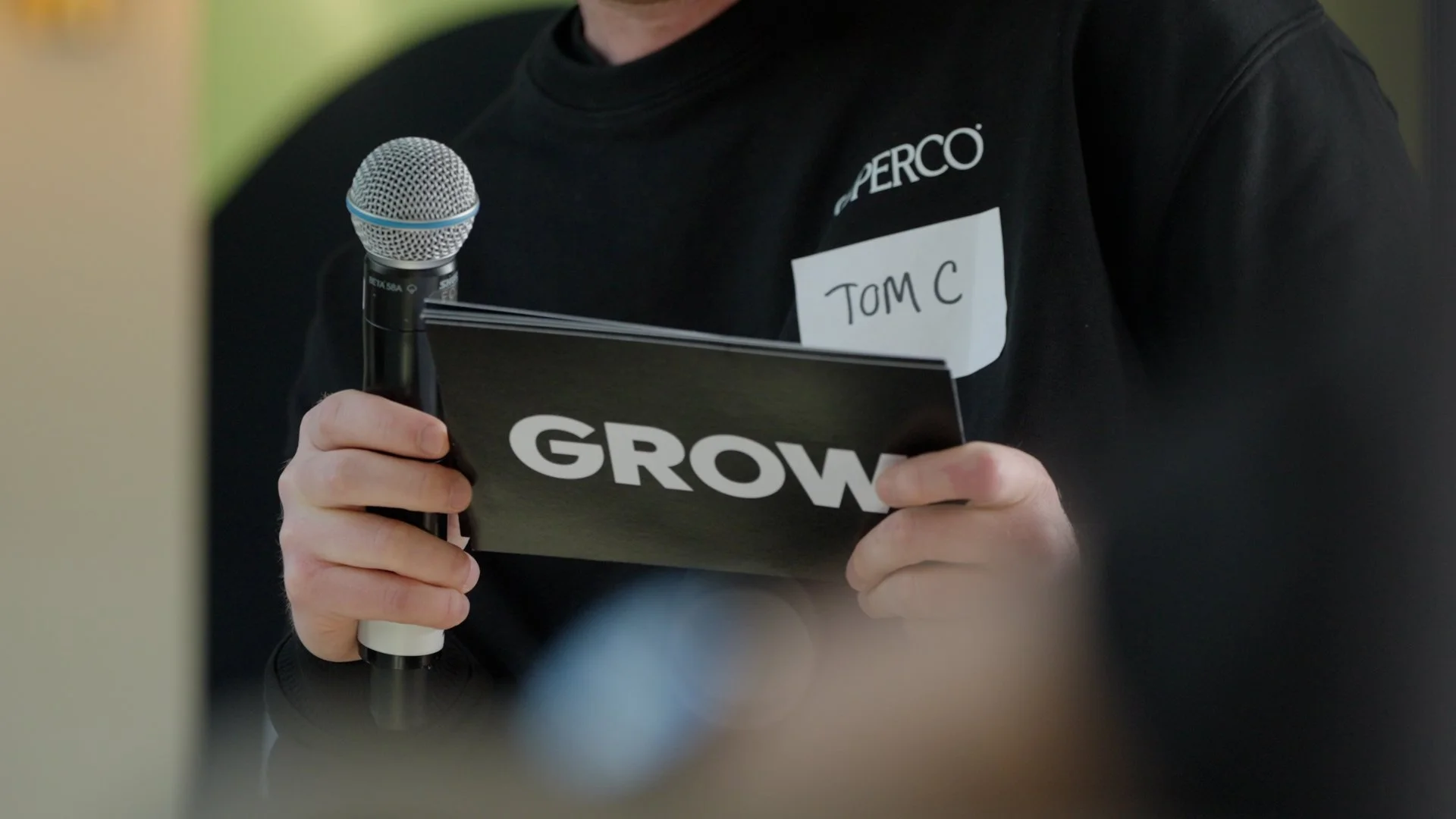 Person holding a microphone and a booklet with the word 'GROW' on it, wearing a black shirt with a name tag reading 'TOM C'.