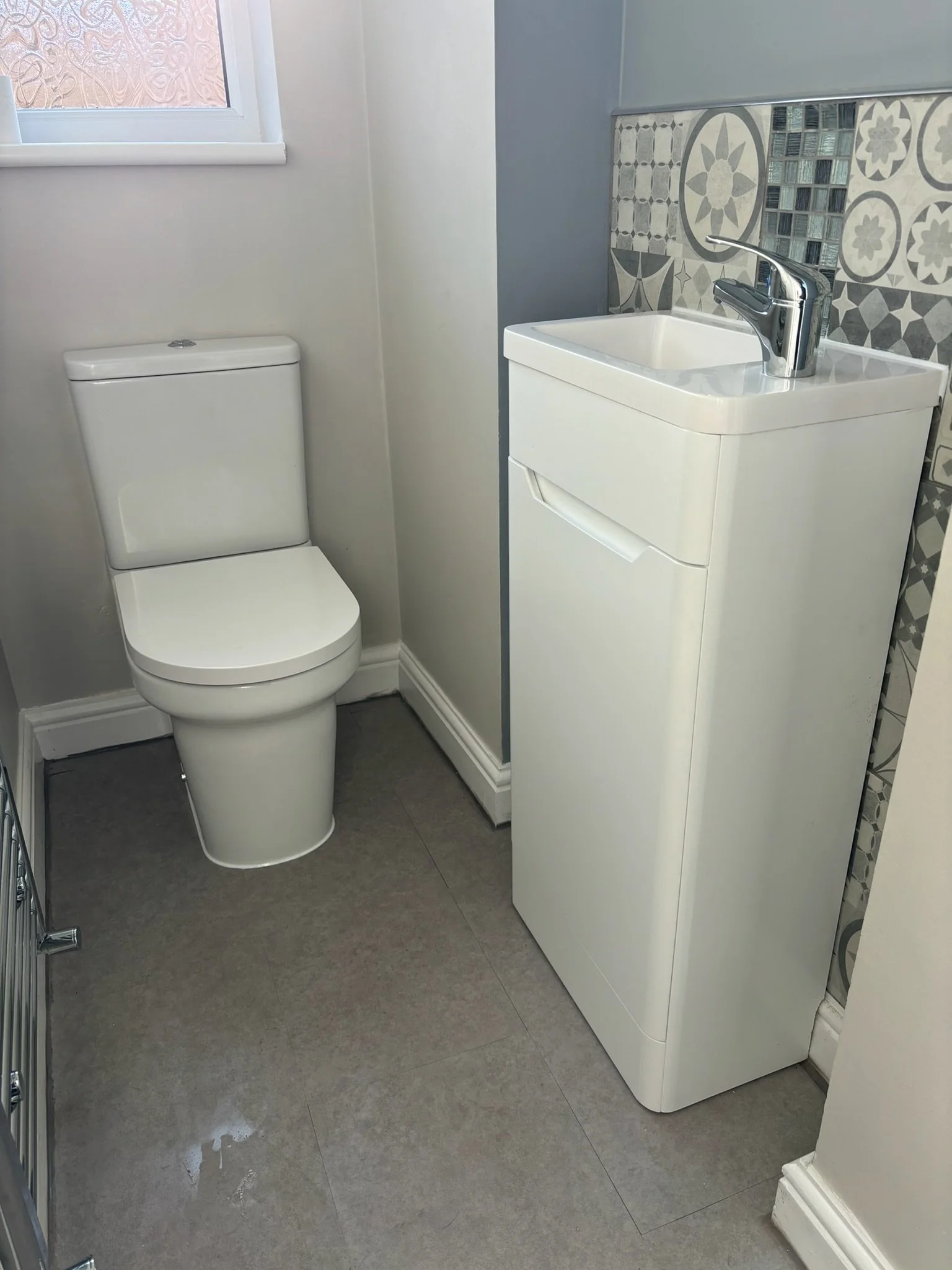 A small bathroom with a toilet and a compact sink unit with a chrome faucet. The wall behind the sink has decorative tile and the floor appears to be tiled. There is a window with frosted glass to the left.