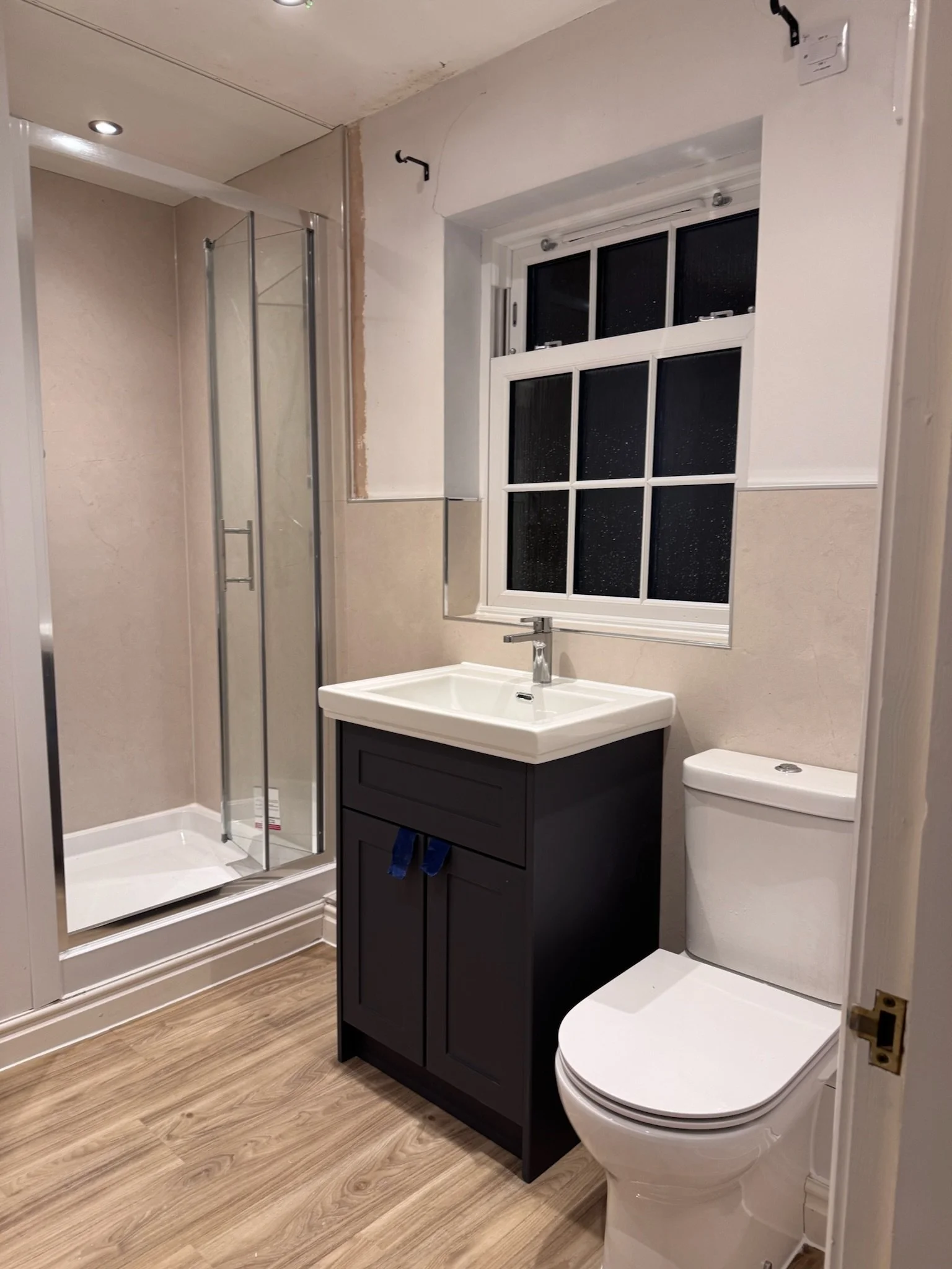 A bathroom with a walk-in shower, a black vanity with a white sink, a toilet, and a window with black panes. The floor is wood, and the walls are partially tiled and painted.