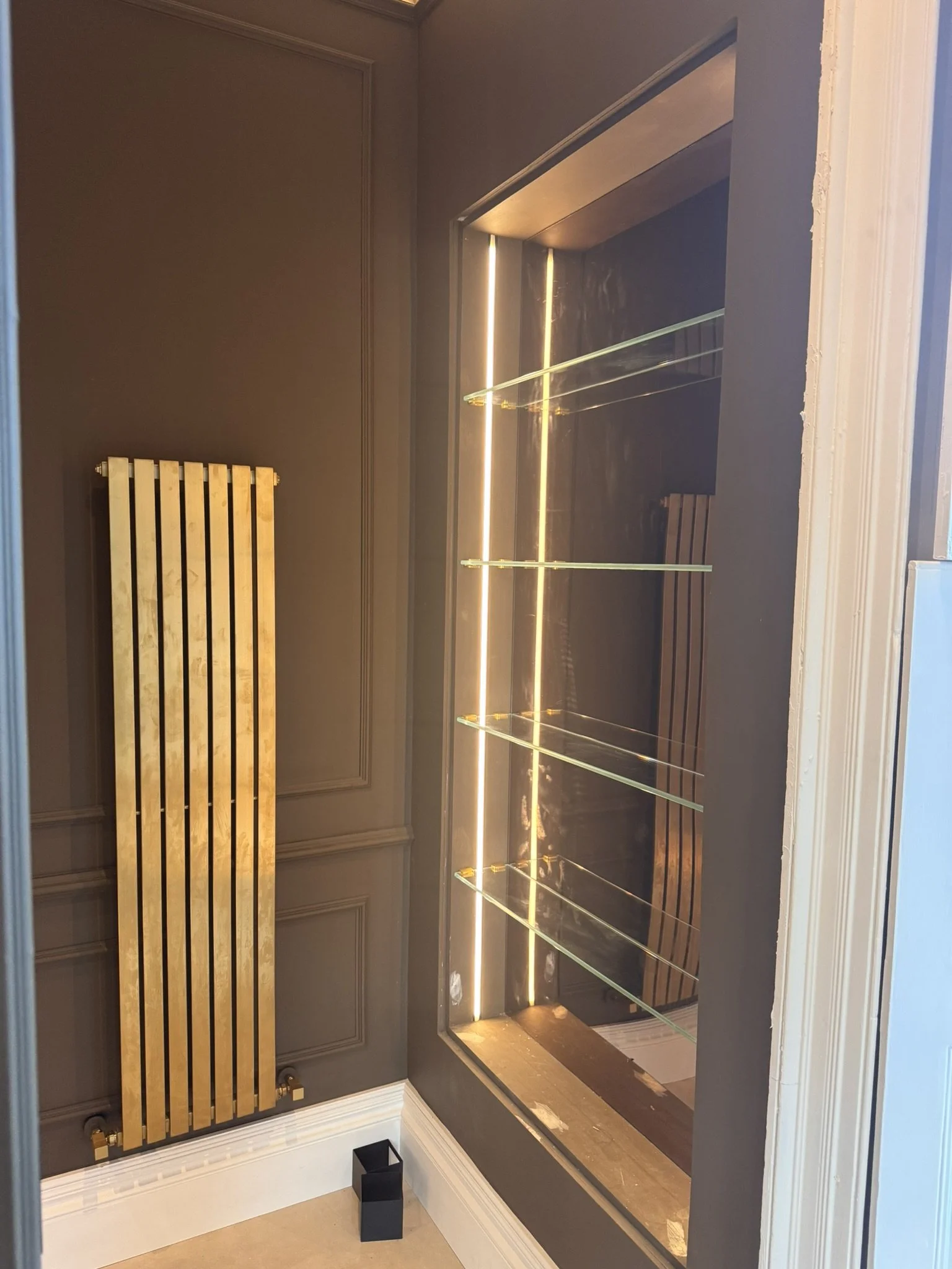 A modern interior space featuring a black wall with a wooden radiator cover, and a glass display case with built-in lighting and multiple glass shelves. The case is set into the wall with a wooden base, and there is a small black trash bin on the flo