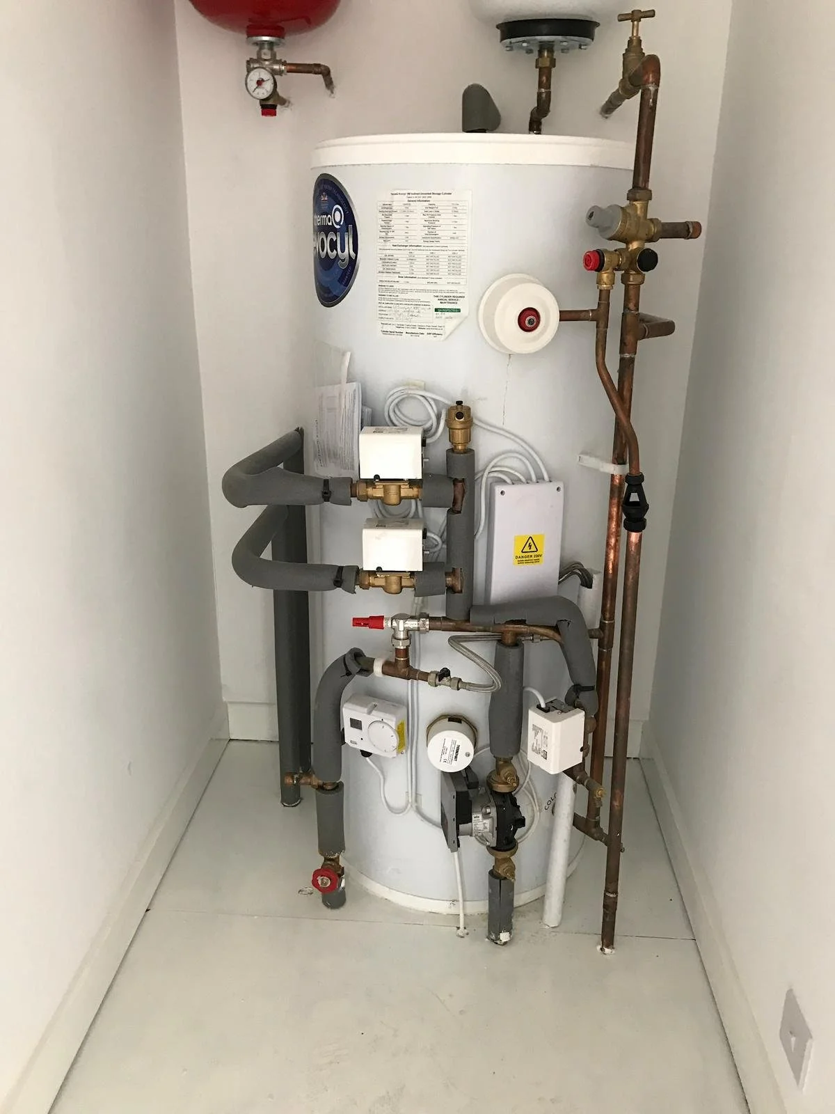 A hot water heater with copper and foam insulation pipes installed on the wall and floor, with pressure and temperature gauges, and shutoff valves mounted in a small utility closet.