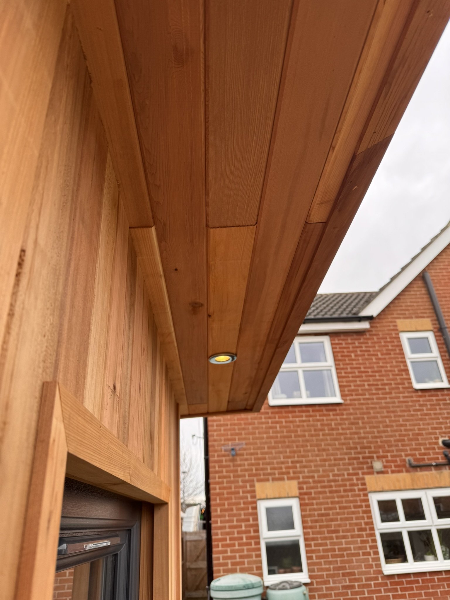 Close-up view of a wooden exterior soffit with a built-in recessed light fixture, adjacent to a brick house with white-framed windows and a cloudy sky in the background.