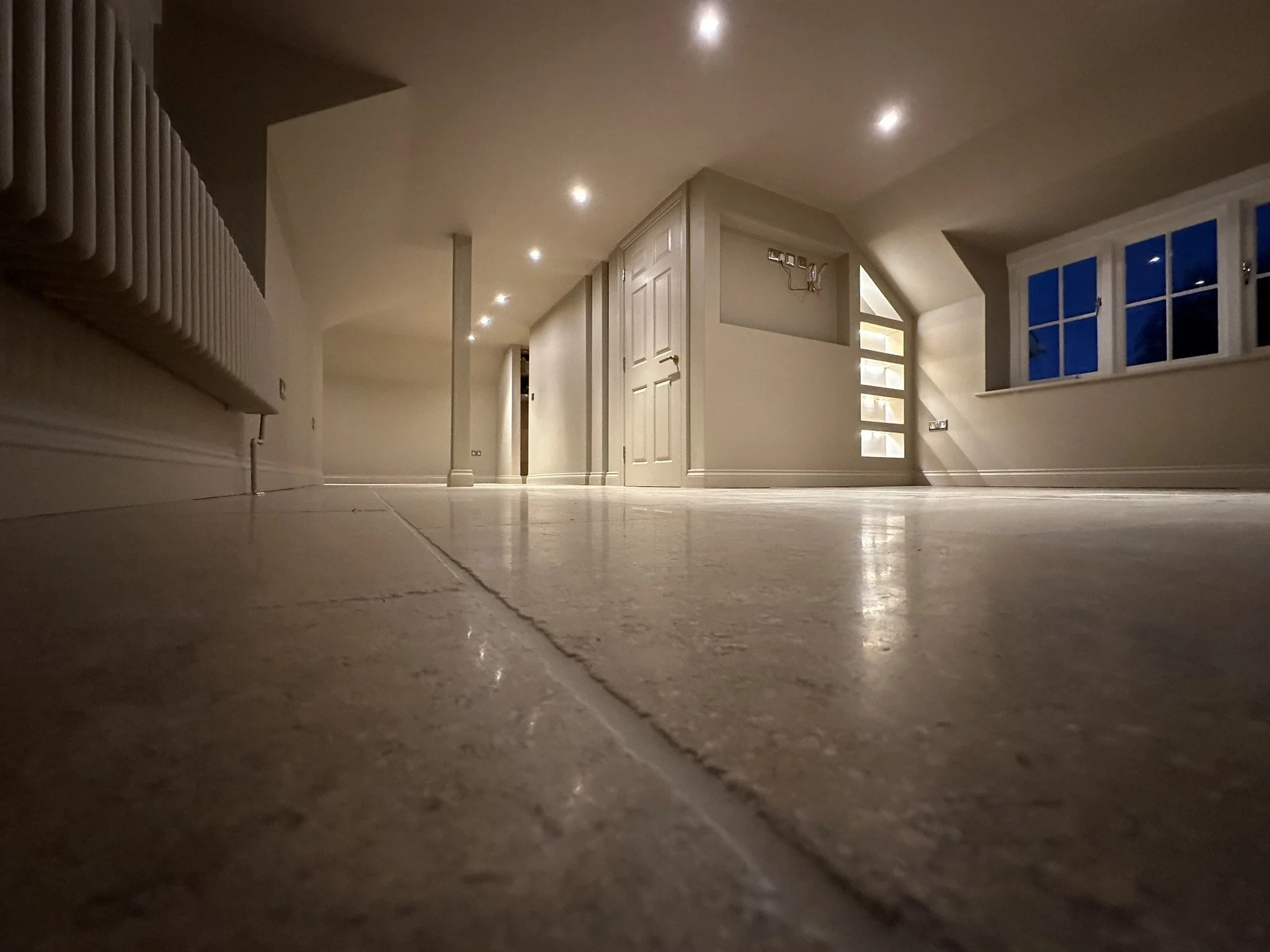 Photo of an empty, well-lit room taken from a low angle, showing a tiled floor, a wall with a radiator on the left, a door and window on the far wall, and ceiling lights.
