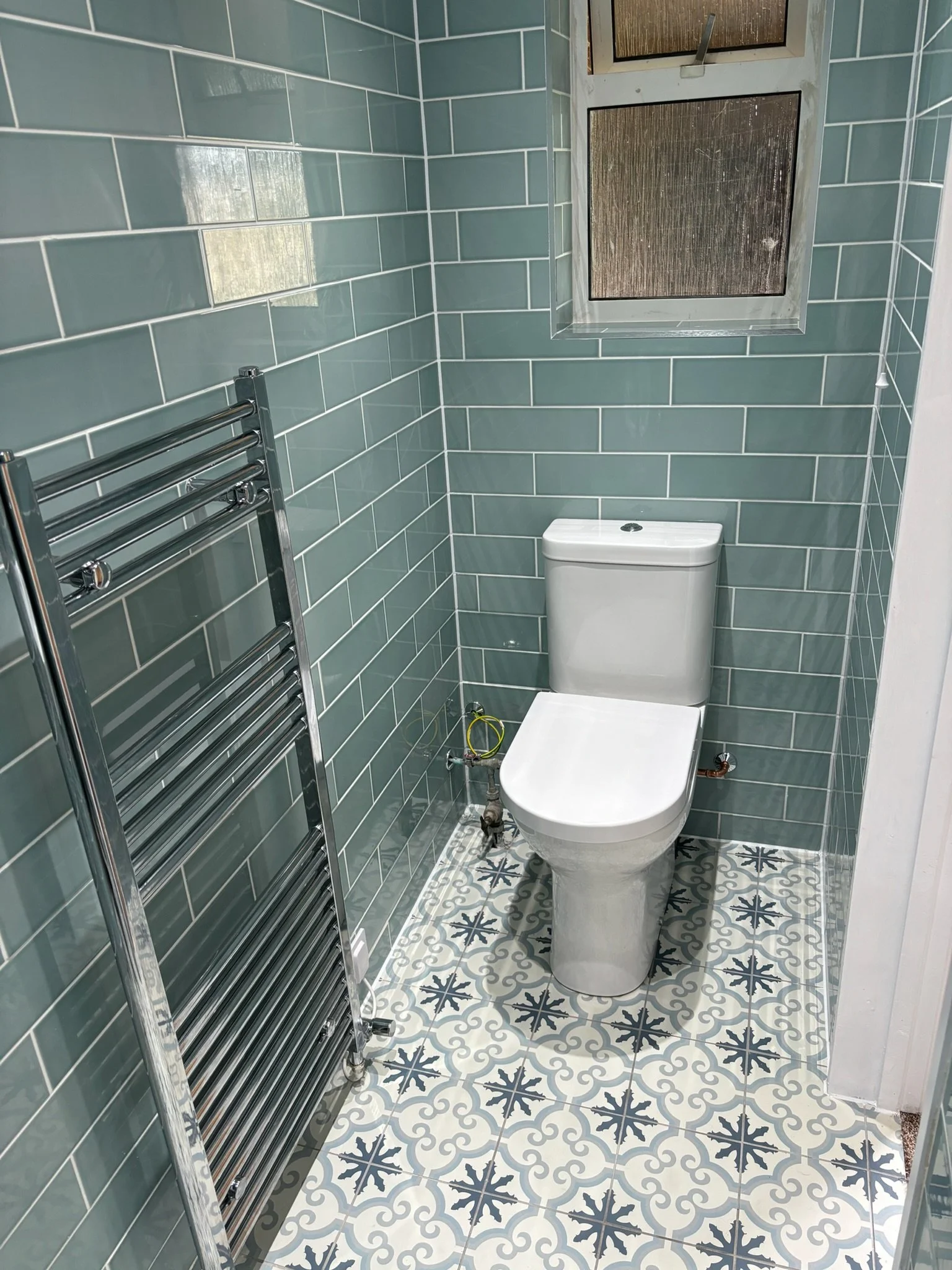 Small bathroom with a white toilet, teal tiled walls, patterned tile floor, a small window, and a chrome towel rack.
