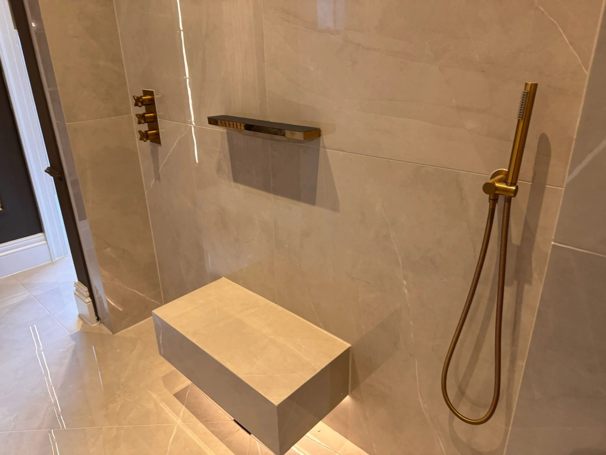 Modern shower area with beige marble tiles, a built-in bench, brass controls, a wall-mounted brass hand shower, and a glass door adjacent to the shower.