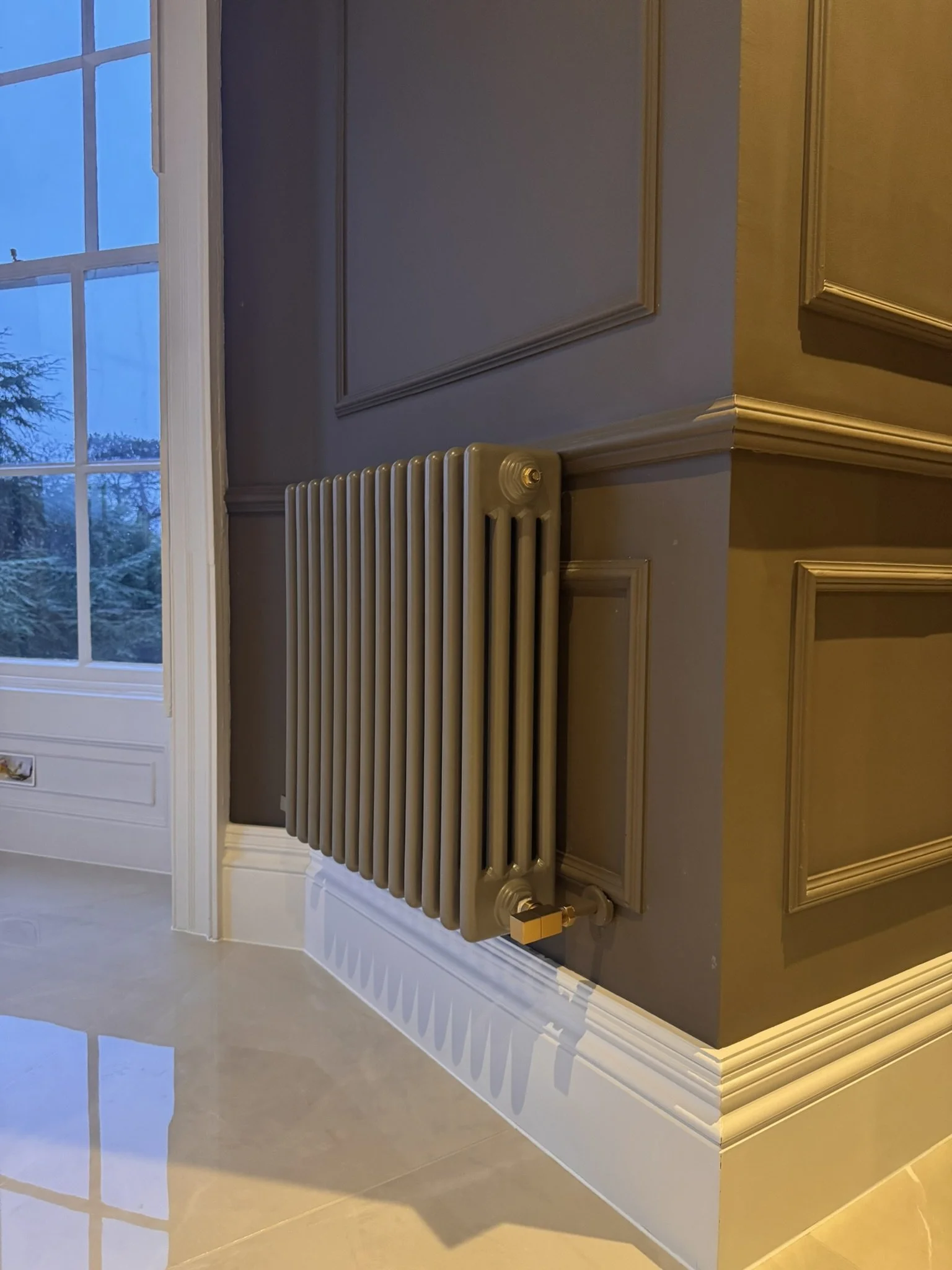 A beige radiator heater mounted on a dark brown wall below decorative molding, with a window to the left showing trees outside and a glossy light-colored tiled floor.