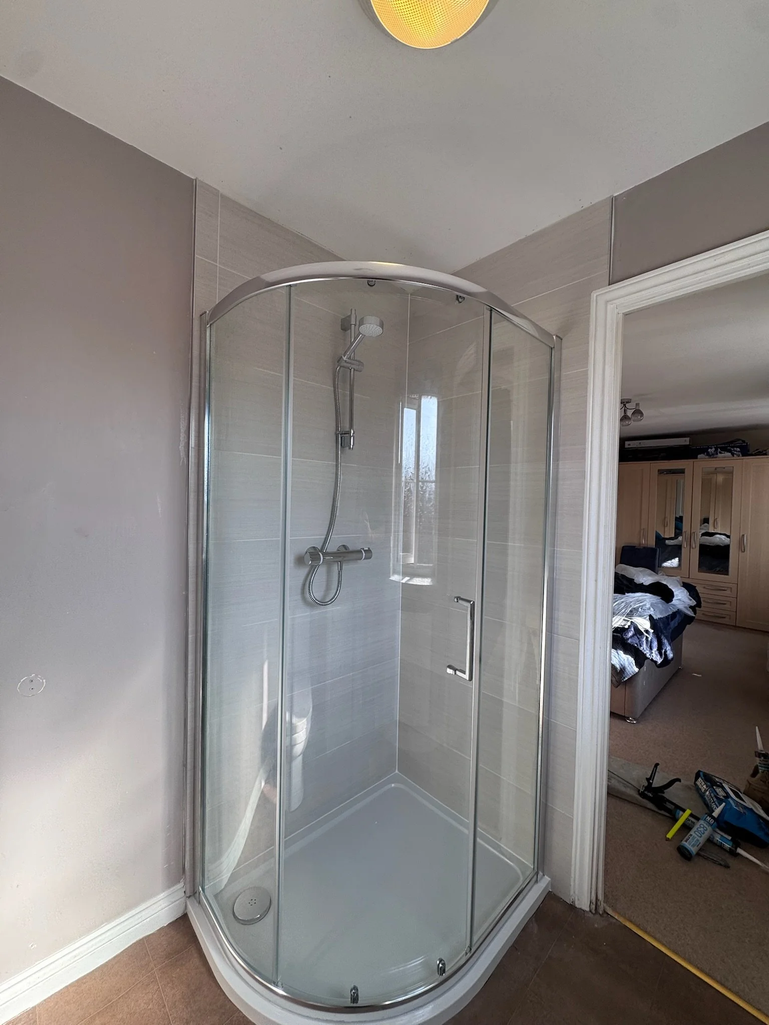 A corner of a bathroom with a glass shower enclosure. The enclosure has a chrome handle and a rainfall showerhead with a handheld shower attached. The shower walls are tiled in a neutral beige color. An open door reveals a bedroom with unmade beds, a