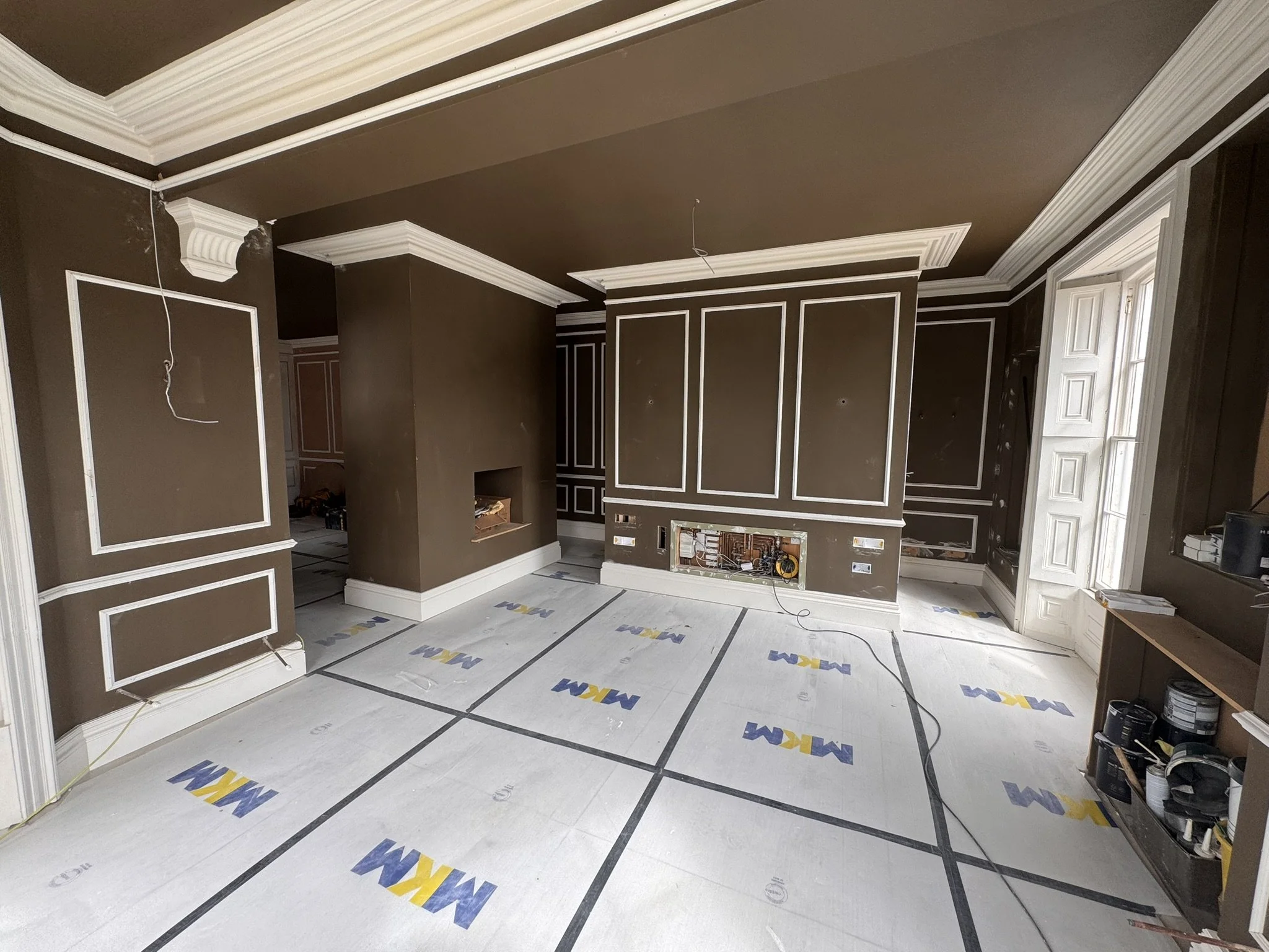 Living room under renovation with brown walls and white crown molding, featuring a large window, a built-in ledge, and construction materials on the floor and shelves.