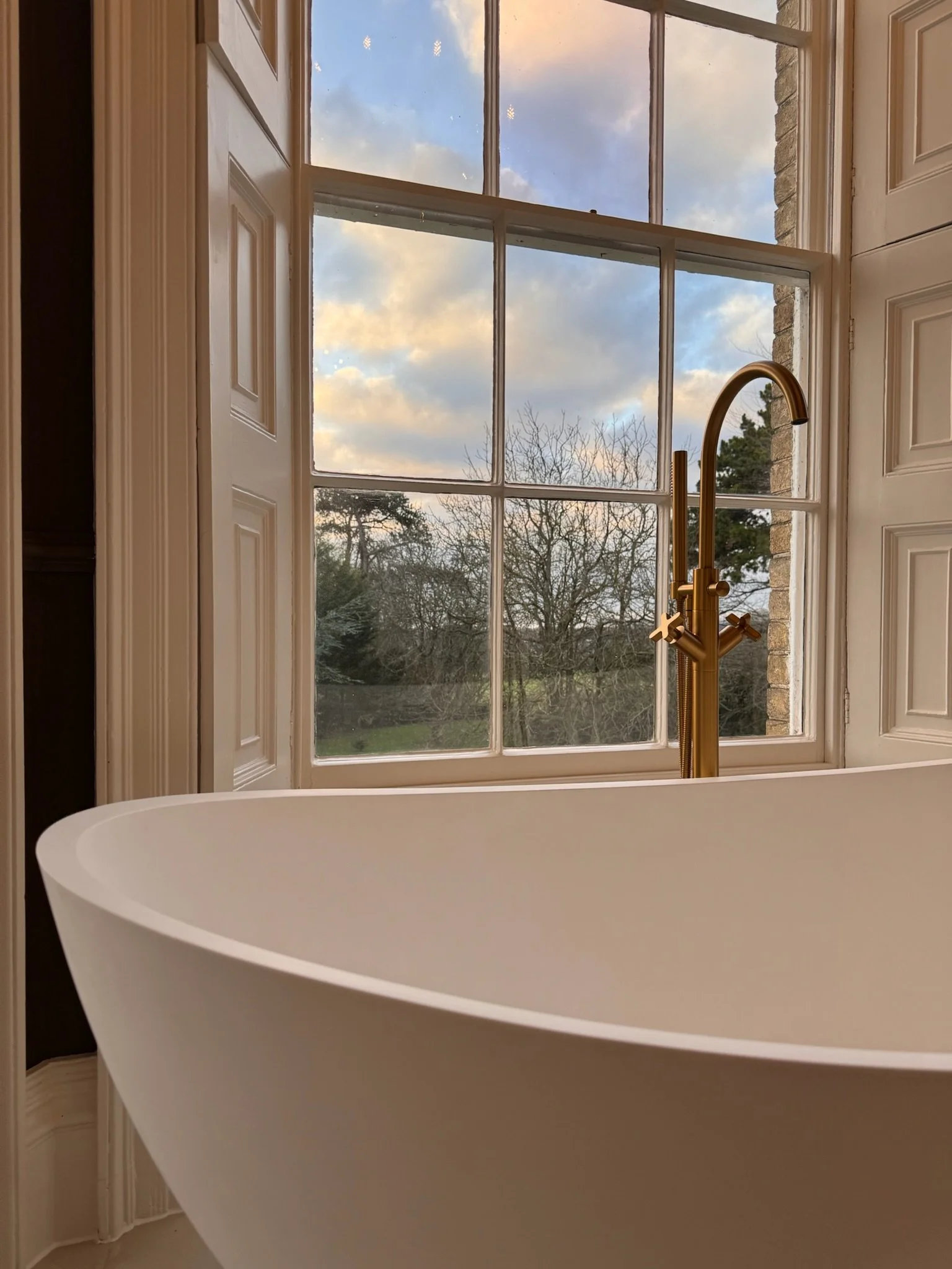 A white freestanding bathtub with a gold faucet is placed in front of a large window with multiple panes, showing a view of trees and a partly cloudy sky.