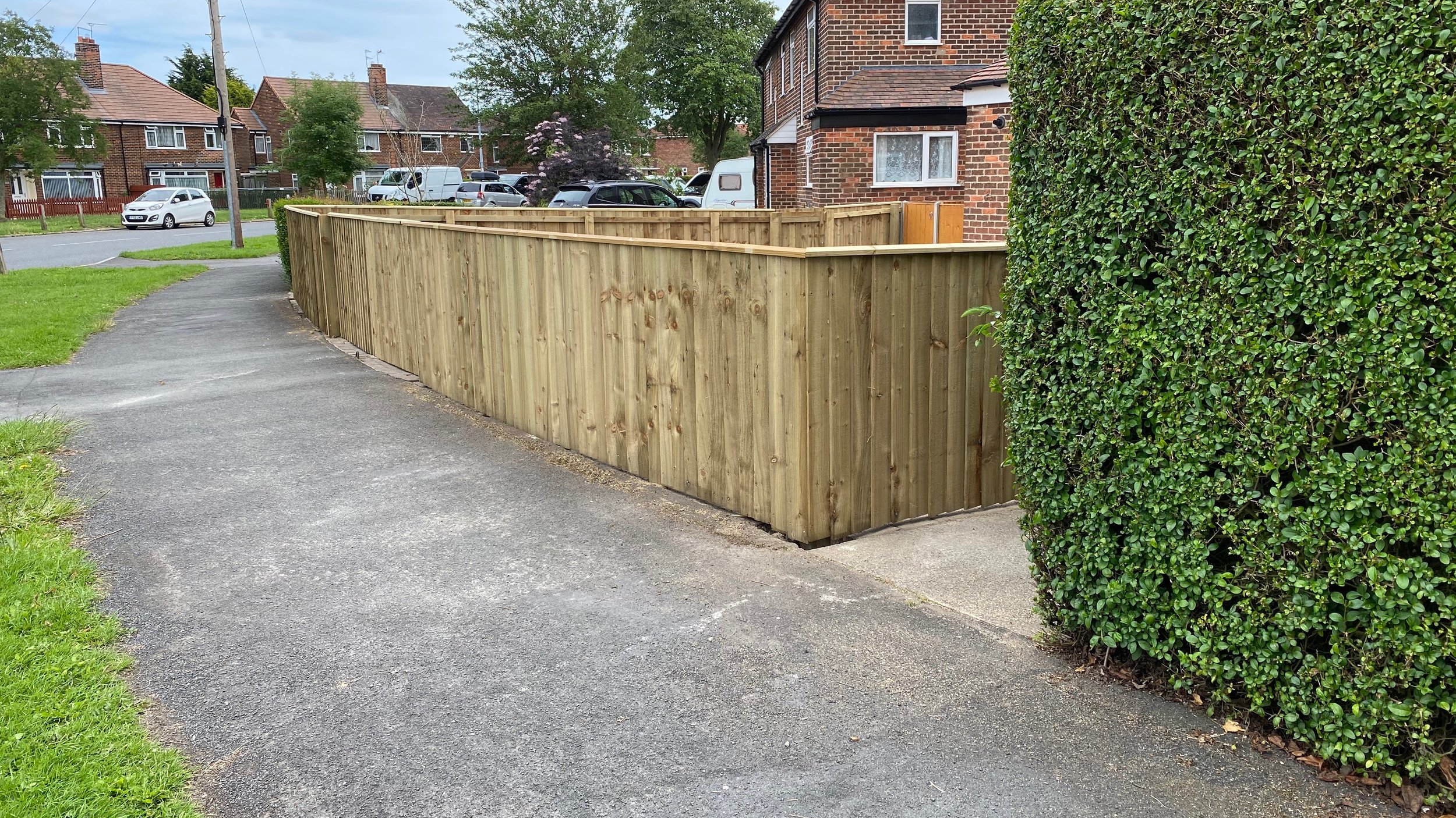 A newly installed wooden privacy fence along a sidewalk in a residential neighborhood, with brick houses, trees, parked cars, and a green hedge nearby.