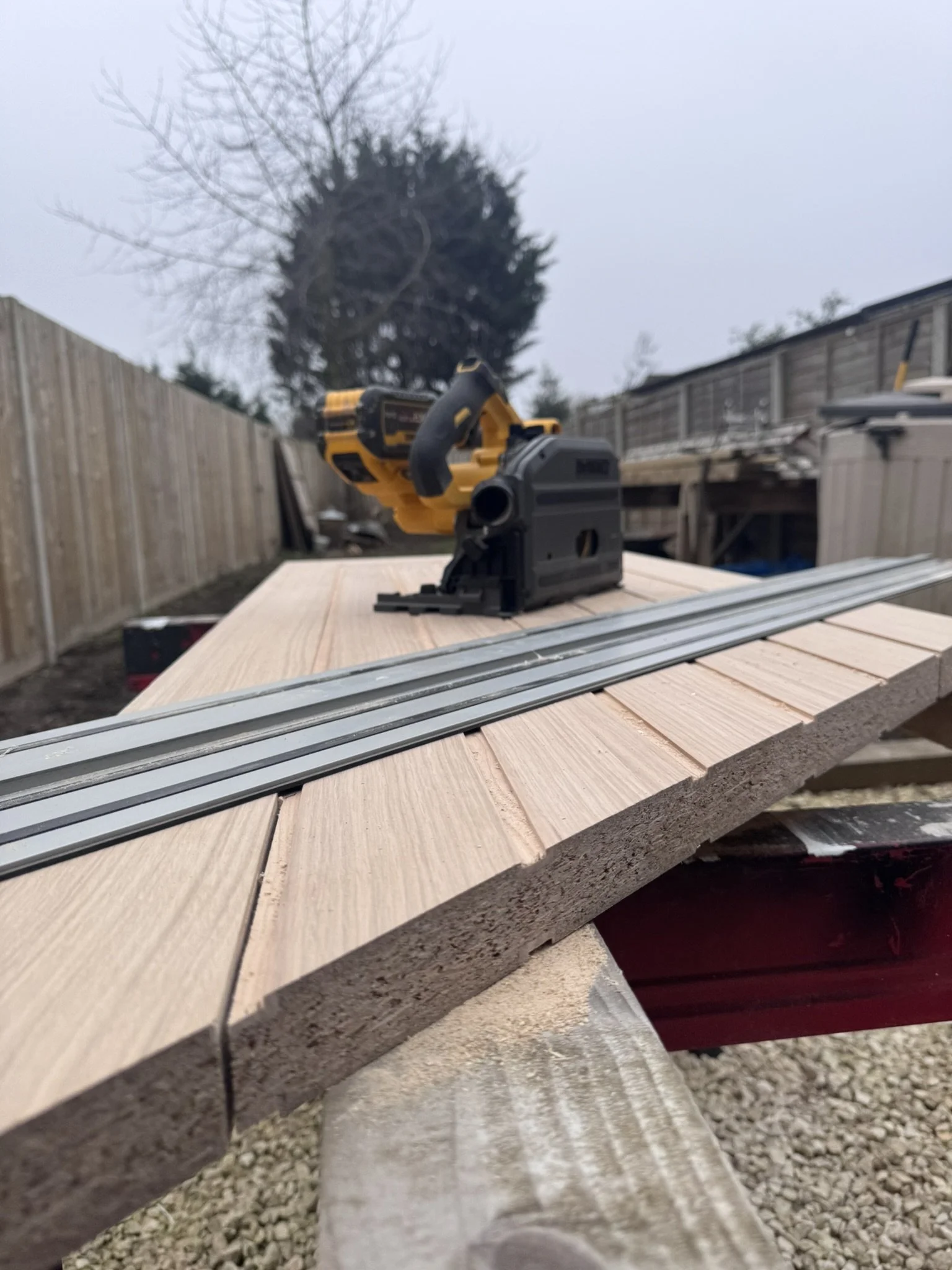 A woodworking project outdoors with a yellow cordless nail gun and metal tracks on a wooden surface, with a background of a wooden fence, trees, and an overcast sky.