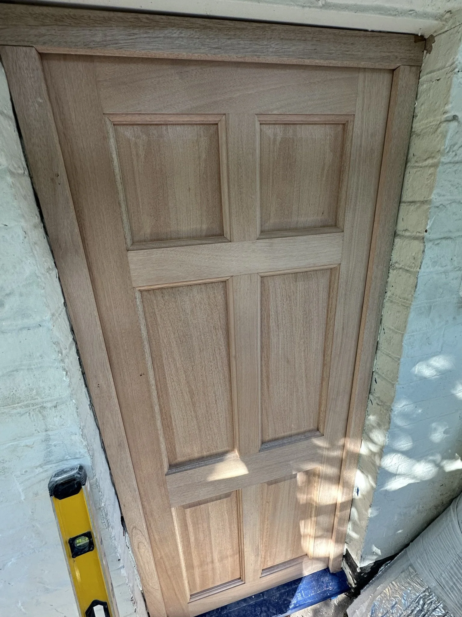 A wooden door with six rectangular panels leaning against a brick wall under construction, with a level tool on the left side and construction materials nearby.