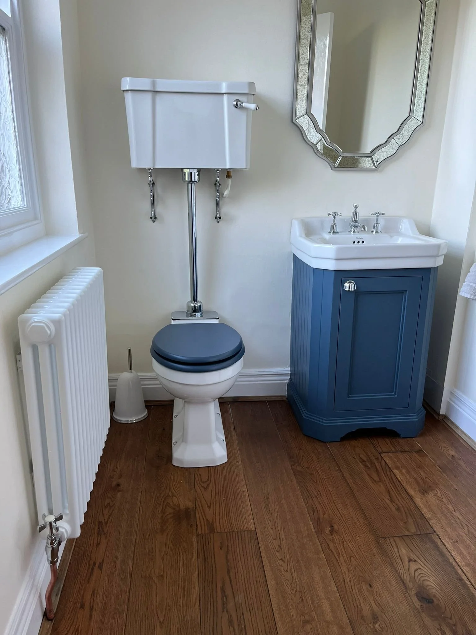A small bathroom with a toilet, a blue sink cabinet, a mirror, and a window with a windowsill. The floor is wooden.