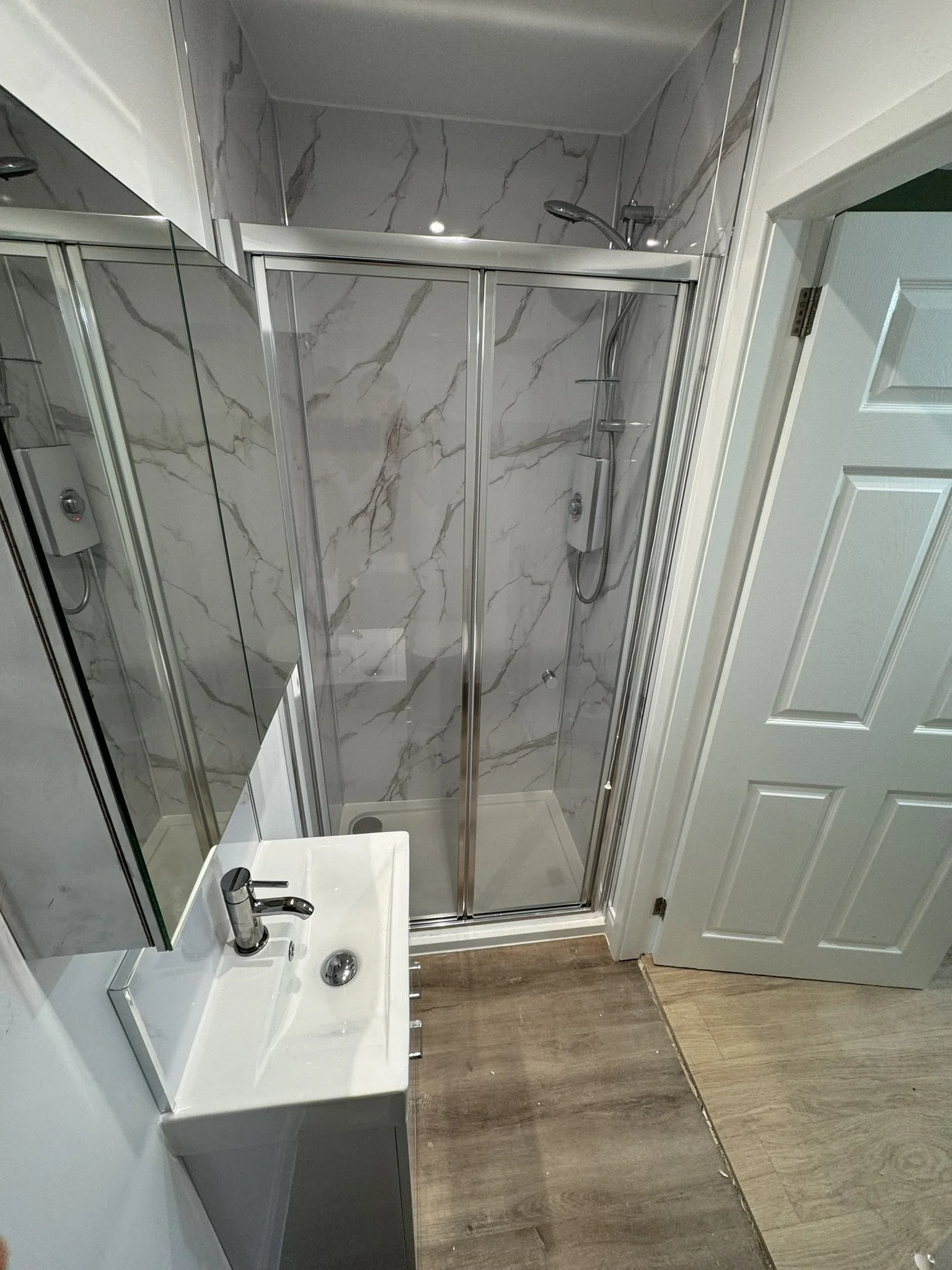 Small bathroom with a glass enclosed shower with marble walls, a small white sink with a modern faucet, mirror, and a wood-patterned floor.