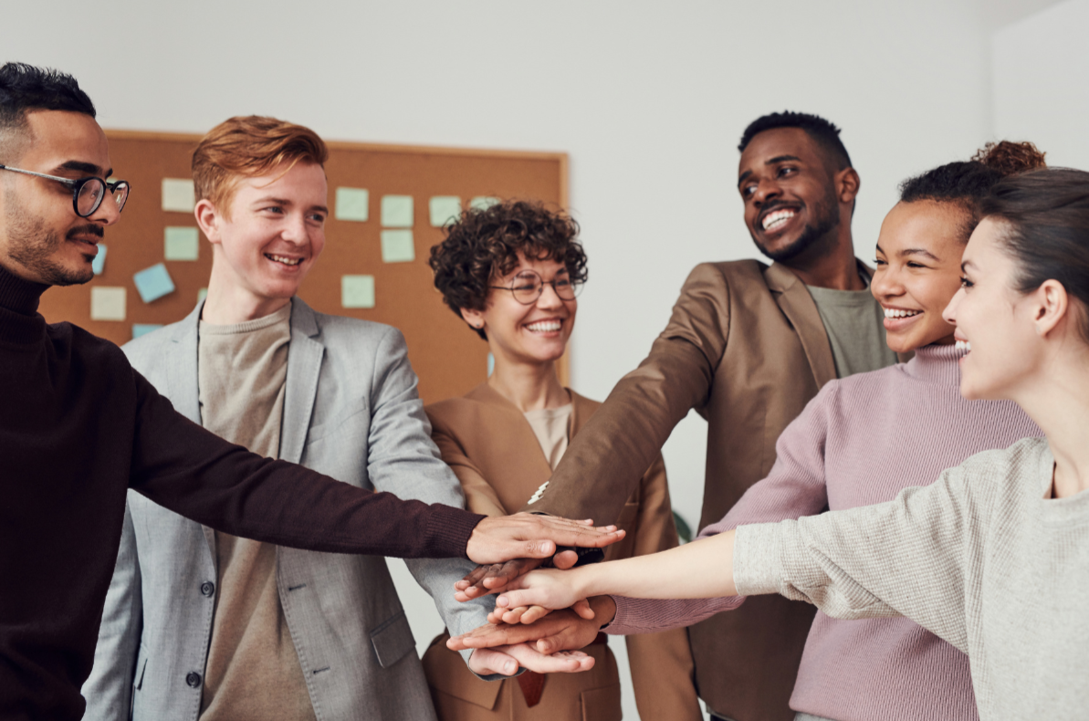 A diverse group of six people in an office, smiling and stacking their hands together in a gesture of teamwork and unity.