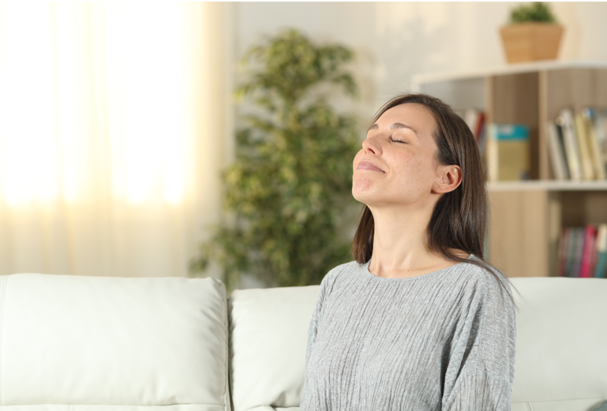 Woman sitting on couch in a bright living room with closed eyes and a peaceful expression.