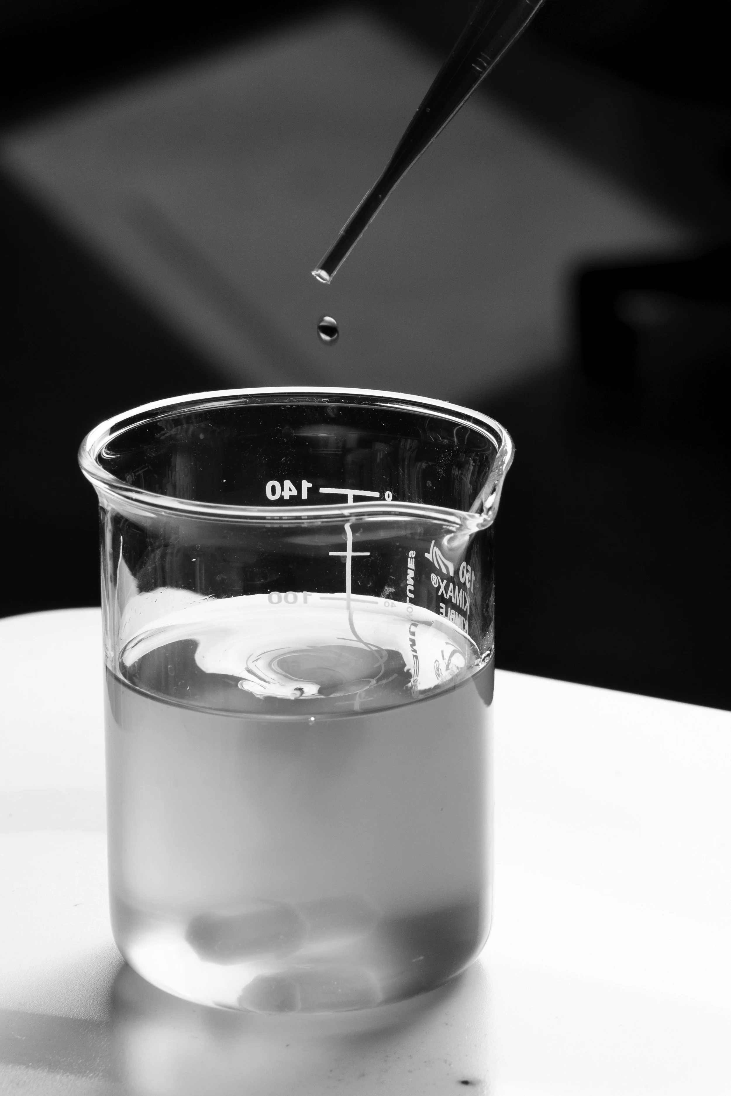 A close-up black-and-white photo of a glass beaker filled with a clear liquid, with a pipette dropping a droplet into it.