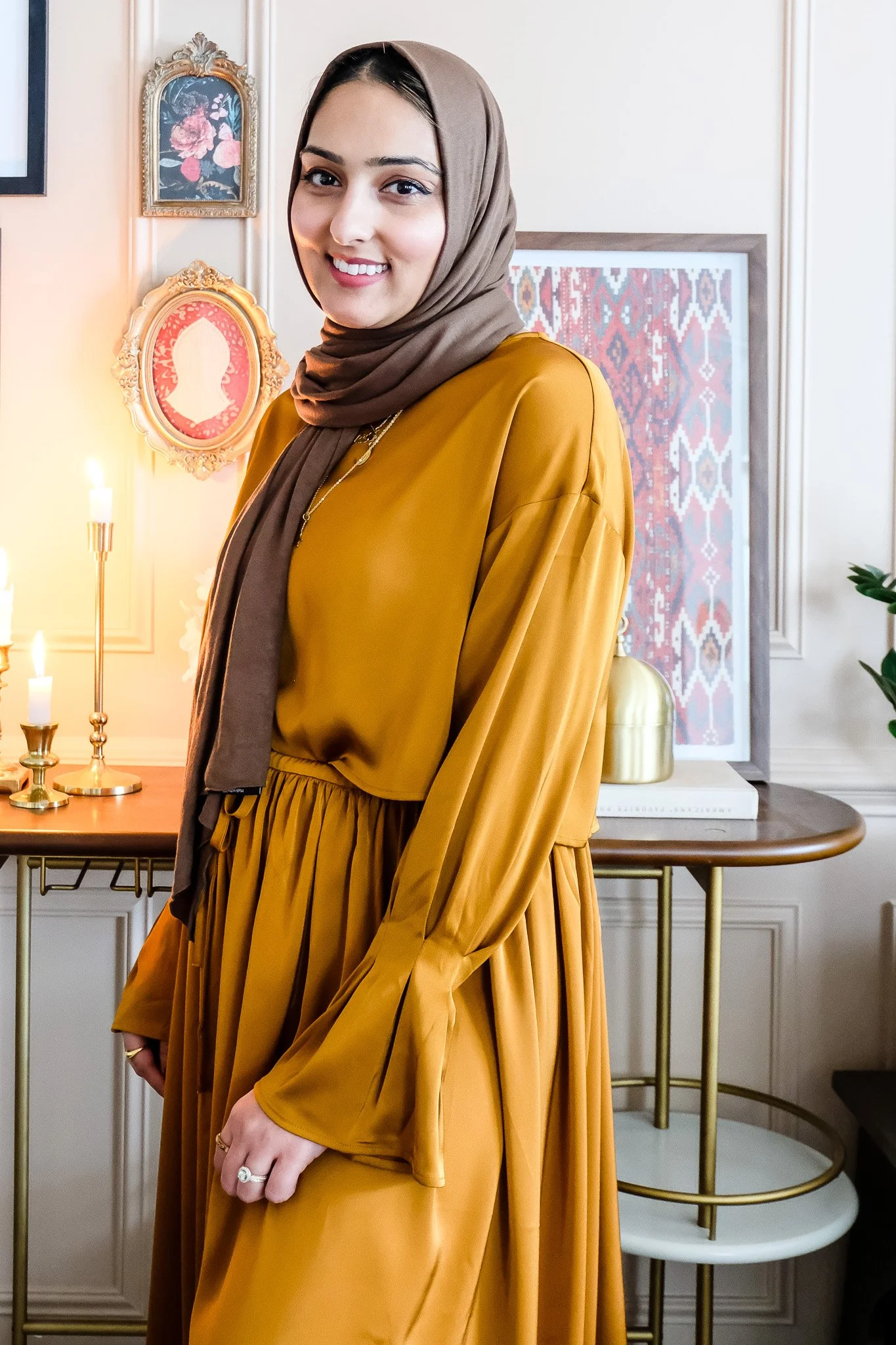 A young woman wearing a brown hijab and a mustard yellow dress, standing indoors in front of a decorative wall with framed artwork and candles.