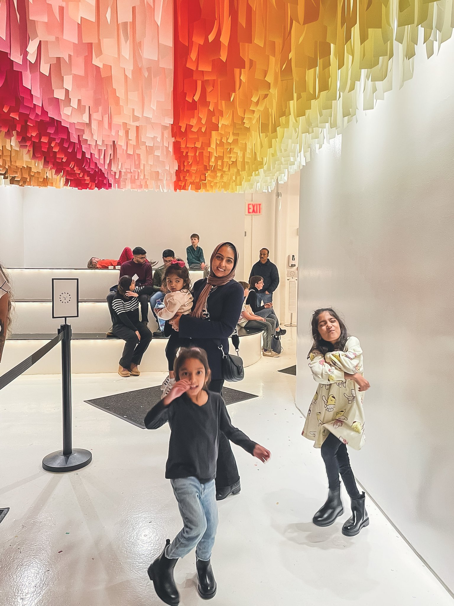A group of children and a woman inside an art gallery. The ceiling has colorful hanging paper strips in a gradient from pink to yellow. The woman is smiling, holding a child, while two young girls pose near the wall, wearing black boots and casual clothes.