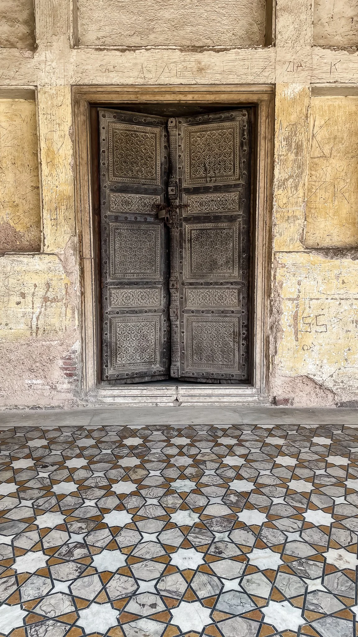 Decorative metal door with intricate patterns, set in a weathered stone or concrete wall with graffiti, and patterned tile flooring in front.